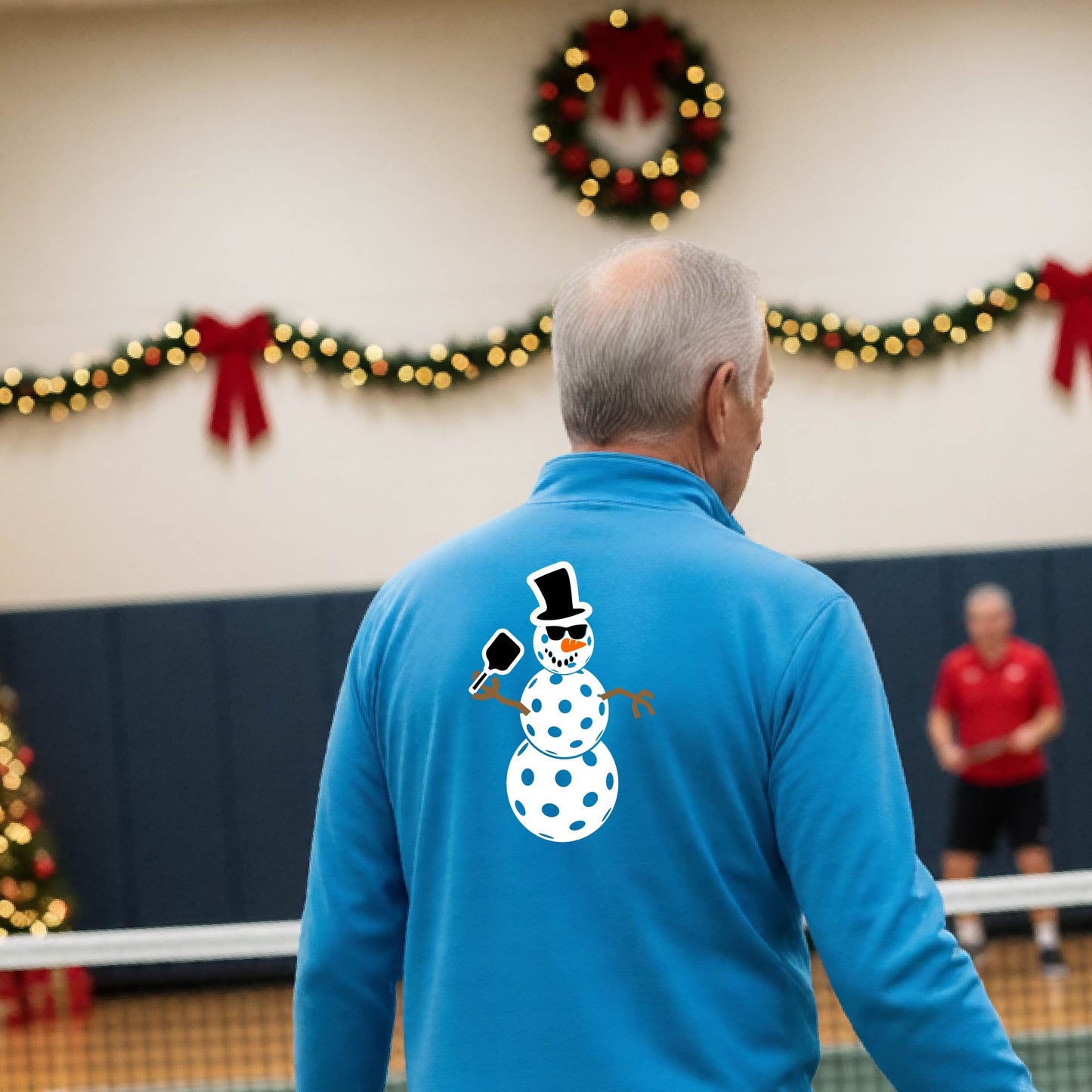 A man viewed from behind wearing a light blue long-sleeve quarter-zip pullover with a white pickleball snowman graphic printed on the back, standing in a decorated pickleball court. 