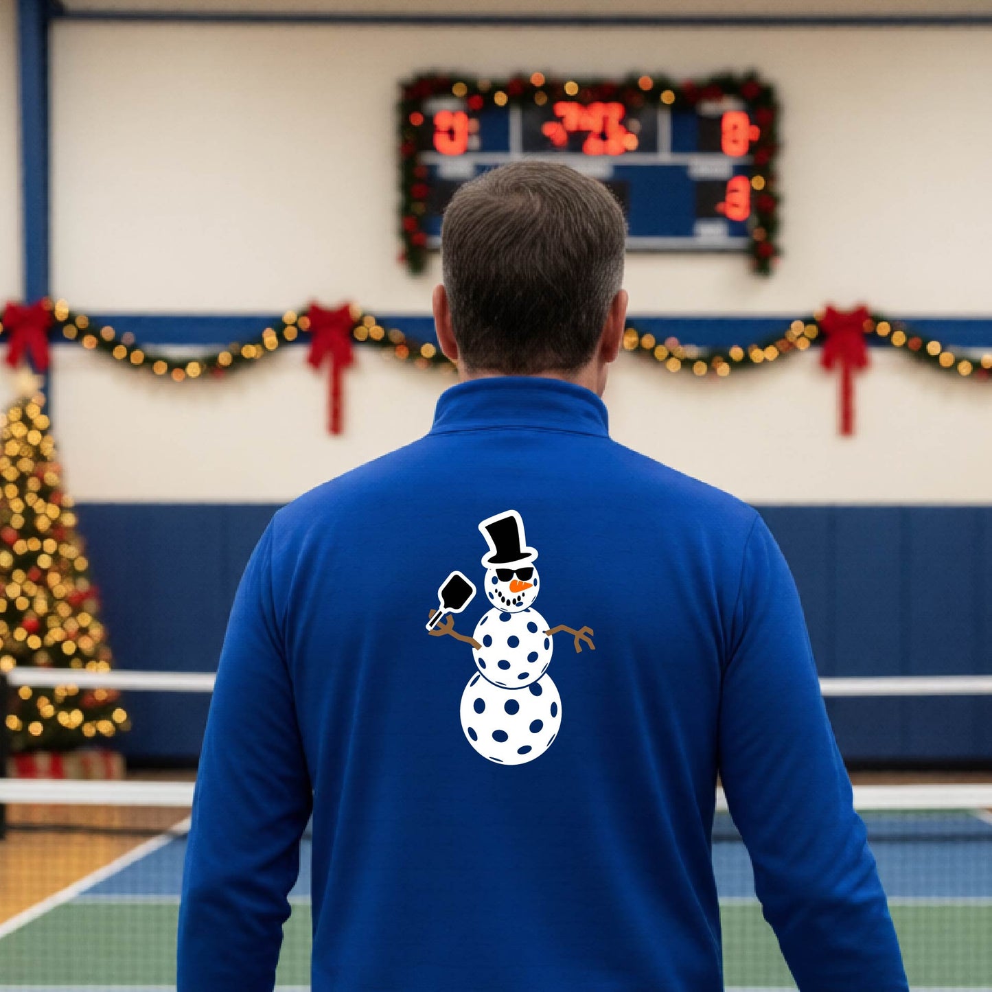A man viewed from behind wearing a royal blue long-sleeve quarter-zip pullover with a white pickleball snowman graphic printed on the back, standing in a decorated pickleball court. 