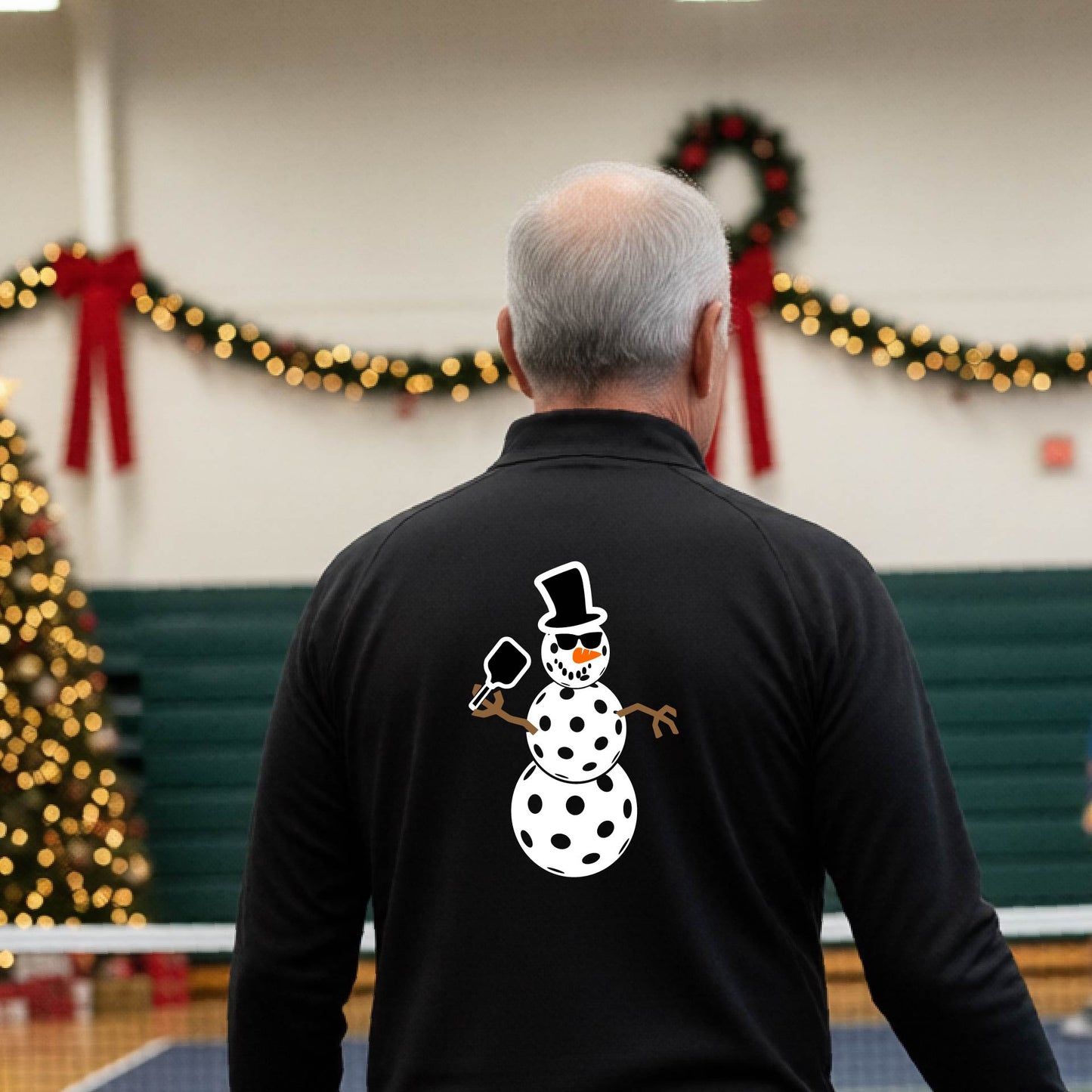 A man viewed from behind wearing a black long-sleeve quarter-zip pullover with a white pickleball snowman graphic printed on the back, standing in a decorated pickleball court. 