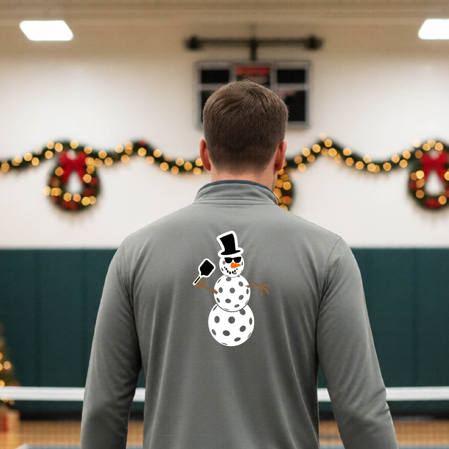 A man viewed from behind wearing a gray long-sleeve quarter-zip pullover with a white pickleball snowman graphic printed on the back, standing in a decorated pickleball court. 