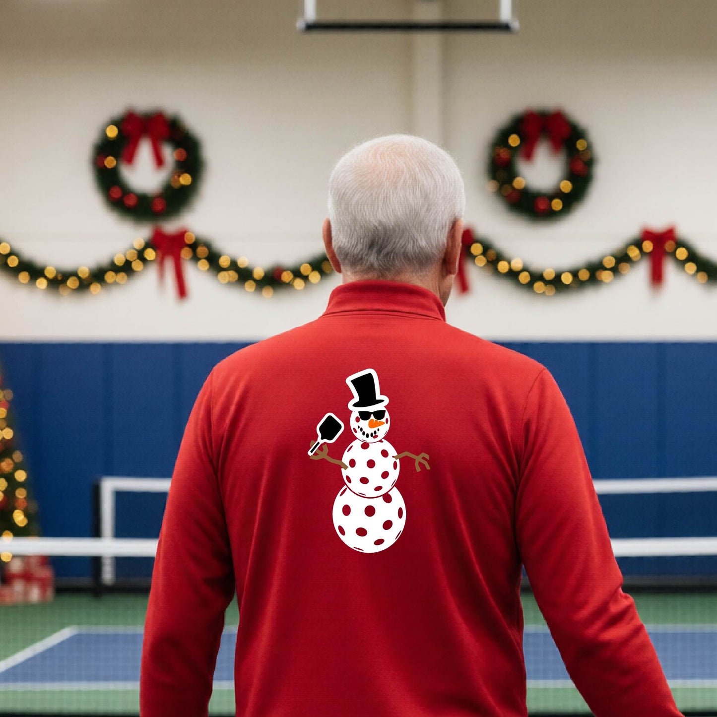 A man viewed from behind wearing a red long-sleeve quarter-zip pullover with a white pickleball snowman graphic printed on the back, standing in a decorated pickleball court. 