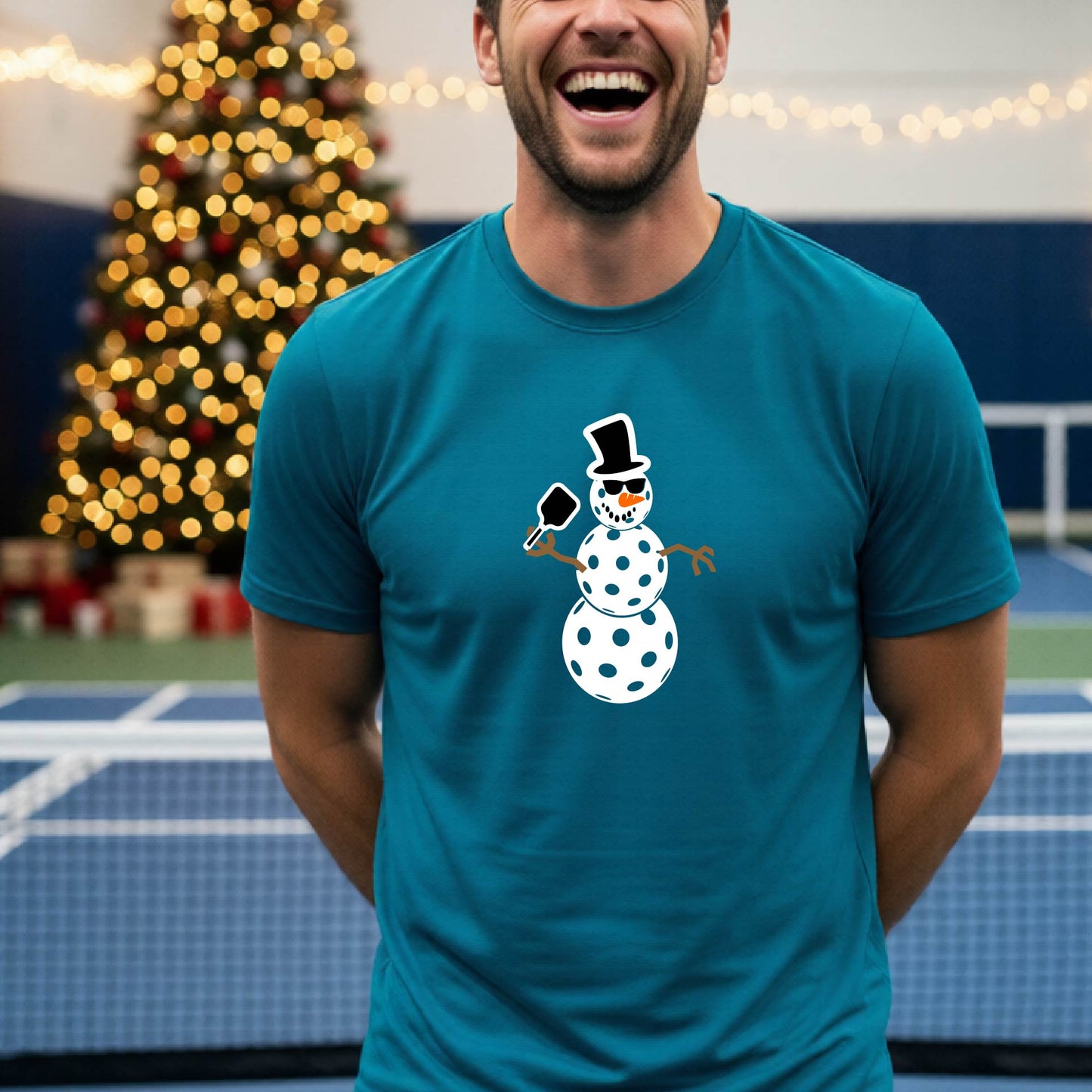 A man wearing a teal blue t-shirt with a white and black pickleball snowman graphic holding a pickleball paddle. The background shows a blurred court with a Christmas tree and string lights.