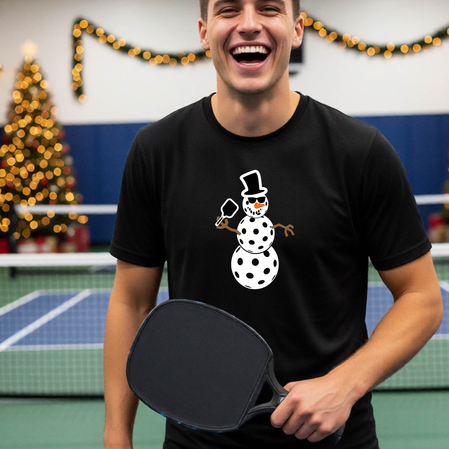 A man wearing a black short-sleeve t-shirt with a white and black pickleball snowman graphic holding a pickleball paddle. The background shows a blurred court with a Christmas tree and string lights.