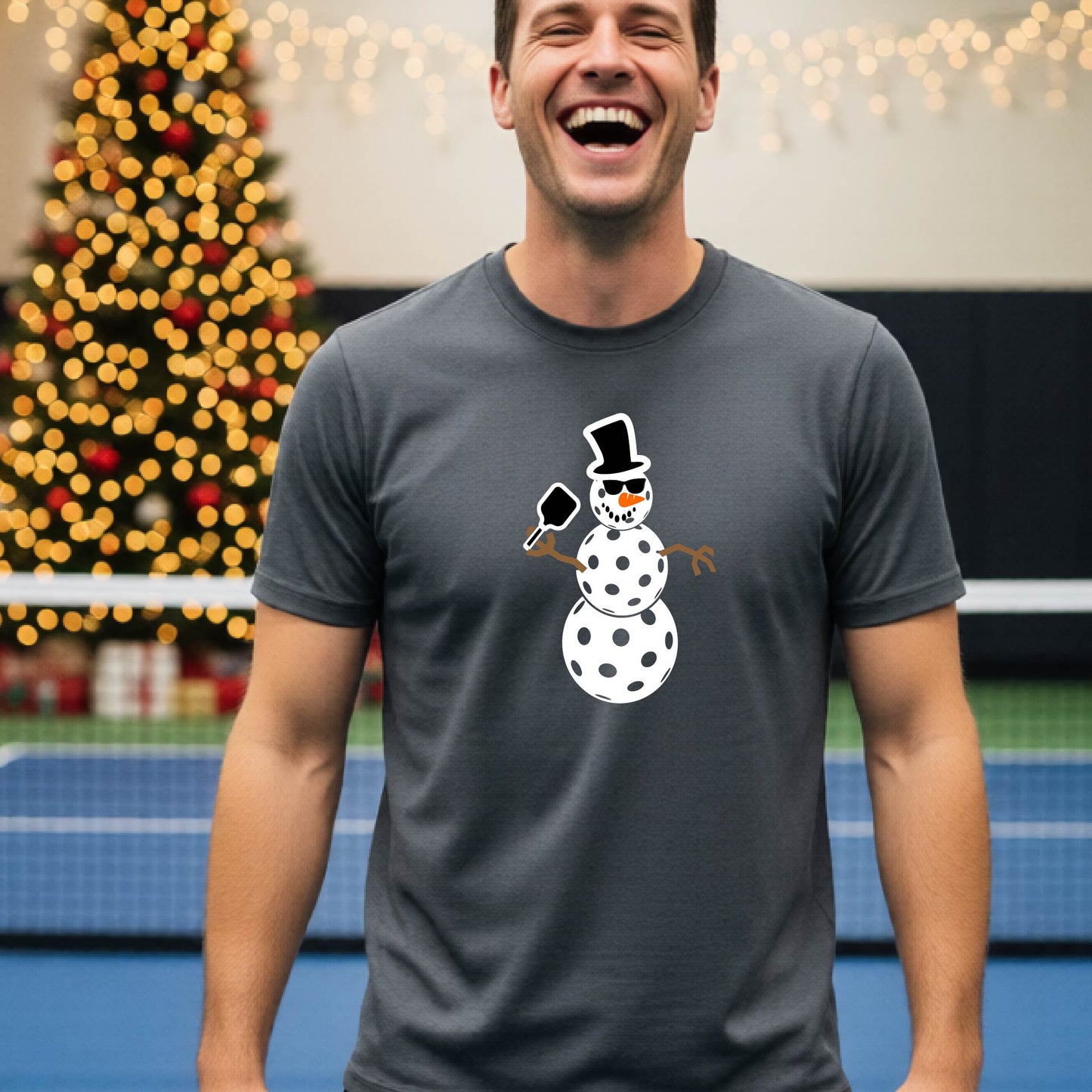 A man wearing a gray short-sleeve t-shirt with a white and black pickleball snowman graphic holding a pickleball paddle. The background shows a blurred court with a Christmas tree and string lights.