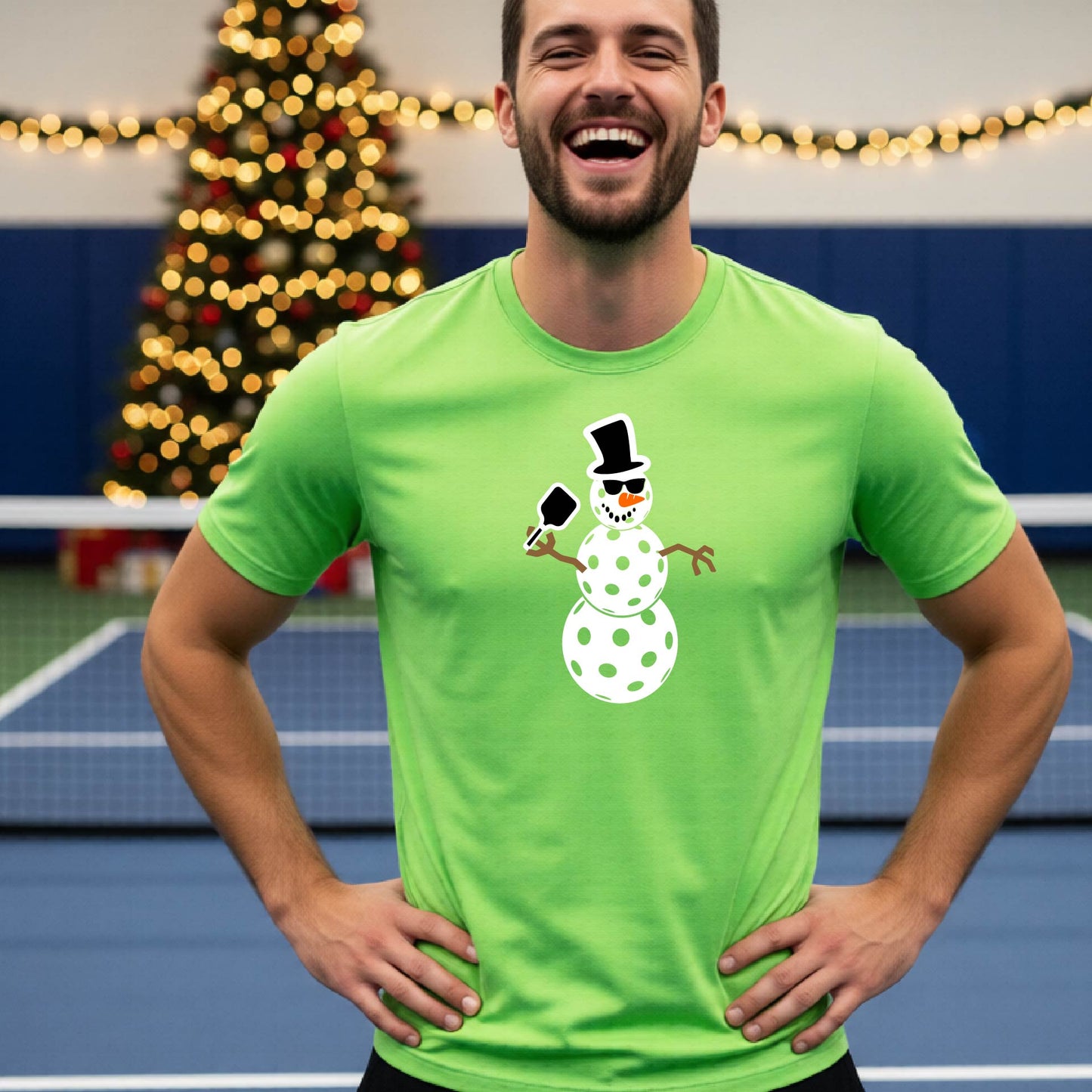 A man wearing a lime green short-sleeve t-shirt with a white and black pickleball snowman graphic holding a pickleball paddle. The background shows a blurred court with a Christmas tree and string lights.