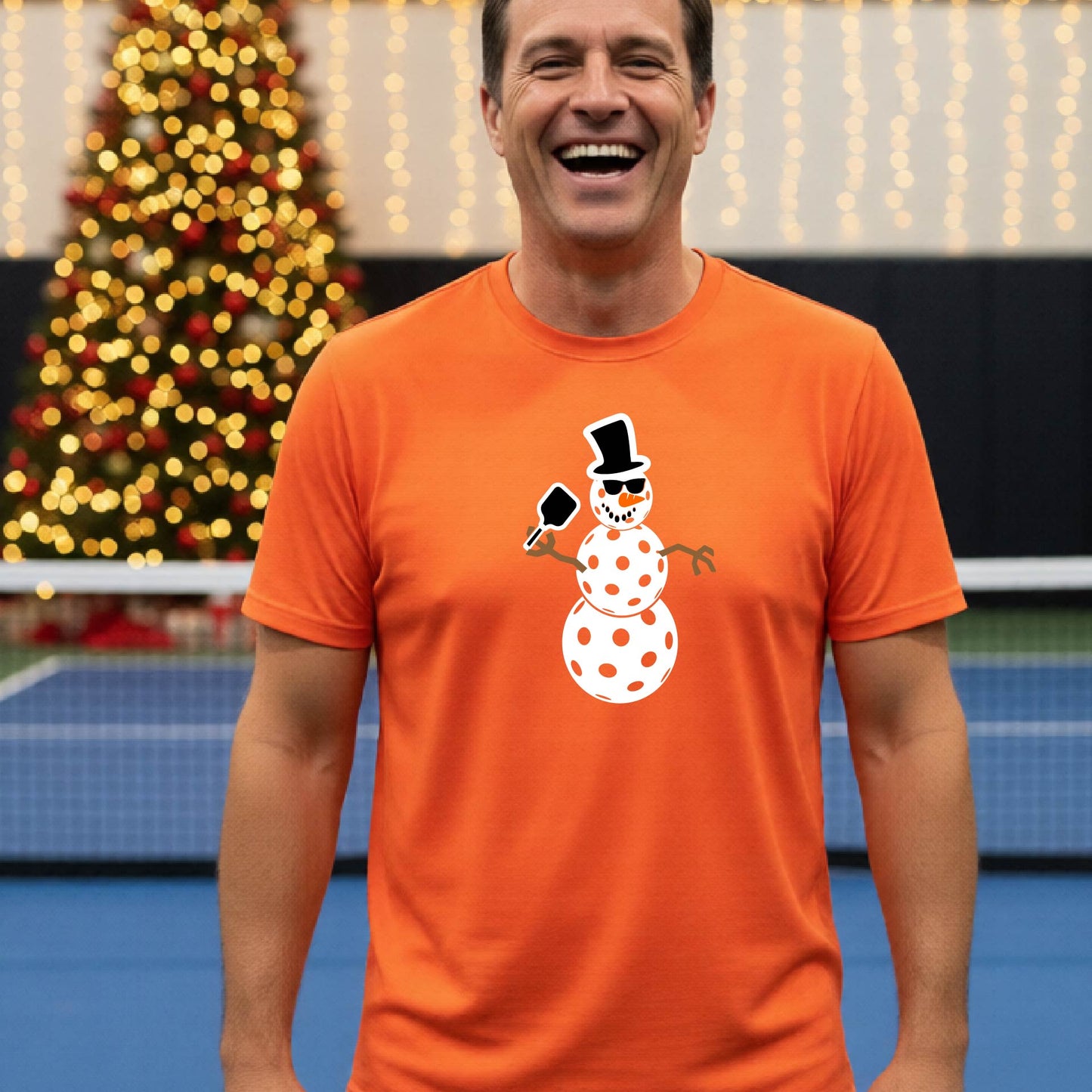 A man wearing a bright orange short-sleeve t-shirt with a white and black pickleball snowman graphic holding a pickleball paddle. The background shows a blurred court with a Christmas tree and string lights.