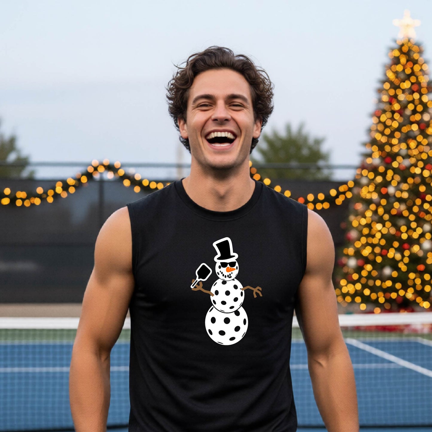 A man wearing a black sleeveless t-shirt with a white and black pickleball snowman graphic holding a pickleball paddle. The background shows a blurred court with a Christmas tree and string lights.