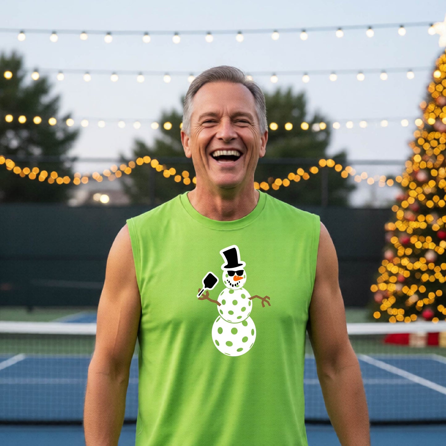 A man wearing a green sleeveless t-shirt with a white and black pickleball snowman graphic holding a pickleball paddle. The background shows a blurred court with a Christmas tree and string lights.