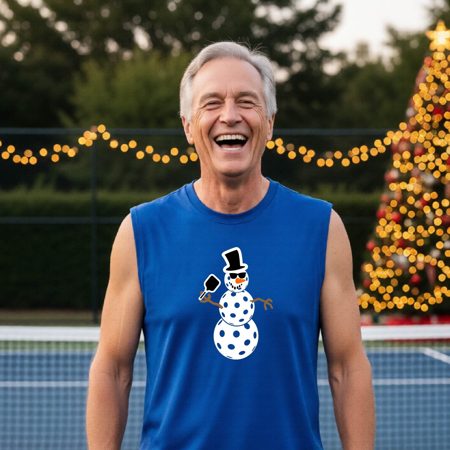 A man wearing a royal blue sleeveless t-shirt with a white and black pickleball snowman graphic holding a pickleball paddle. The background shows a blurred court with a Christmas tree and string lights.
