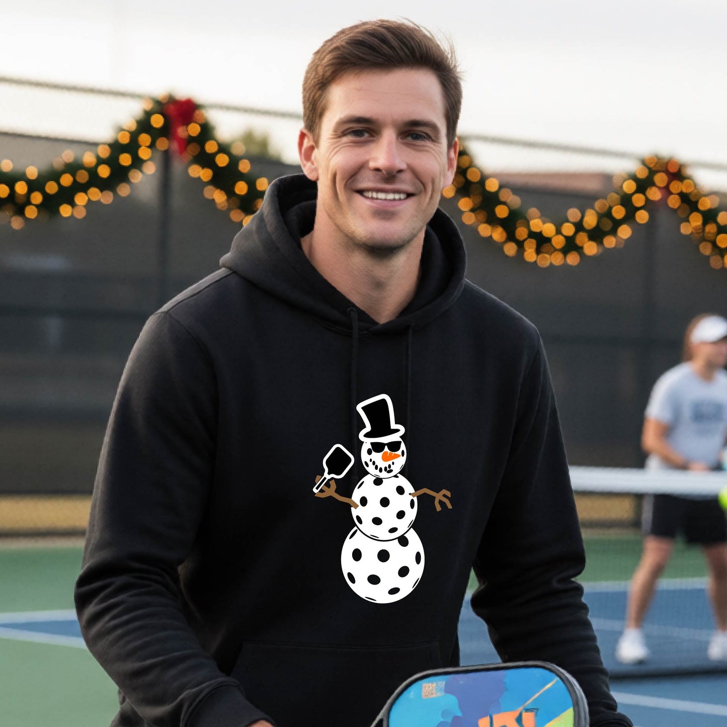 An man smiling on a picleball court wearing a black hoodie with a white and blue pickleball snowman graphic on the front.