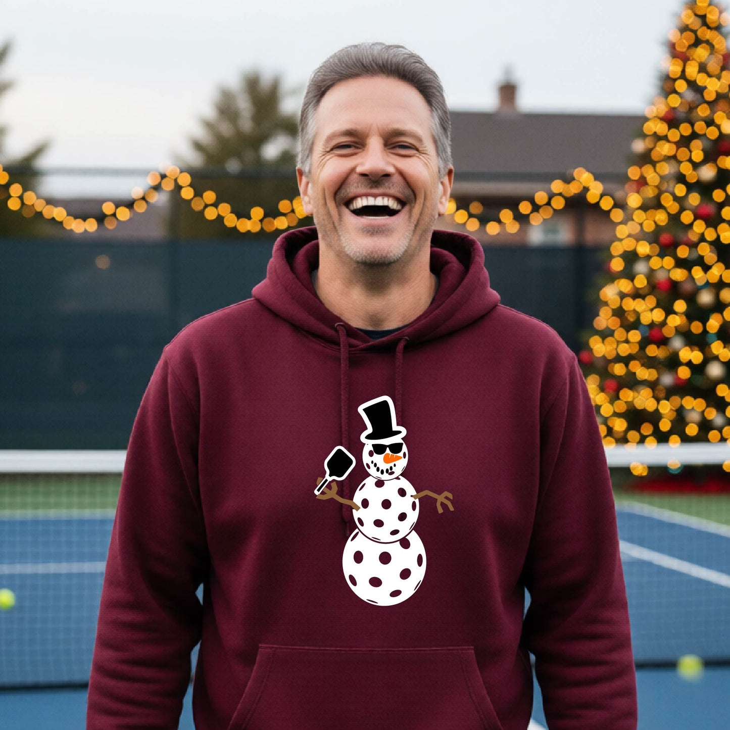 An man smiling on a pickleball court wearing a maroon hoodie with a white and blue pickleball snowman graphic on the front.