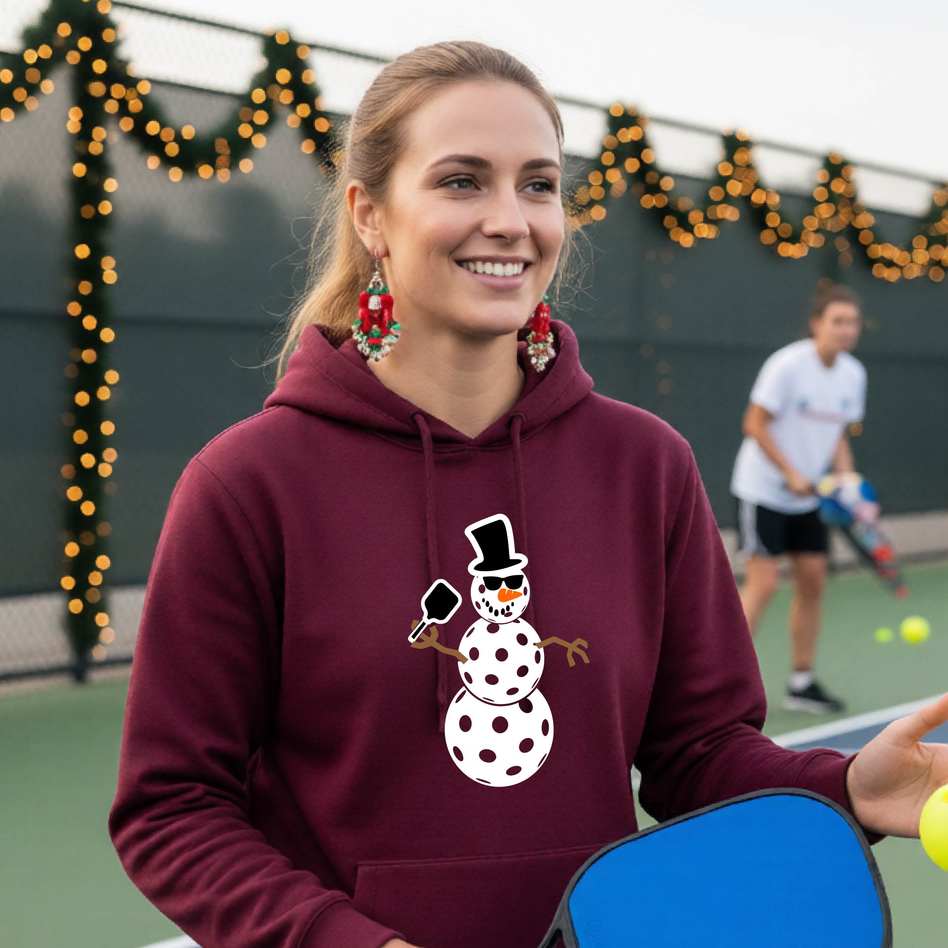 An woman smiling on a picleball court wearing a maroon hoodie with a white and blue pickleball snowman graphic on the front.
