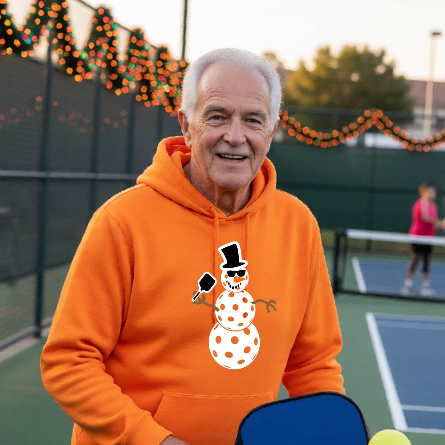 An man smiling on a picleball court wearing a bright orange hoodie with a white and blue pickleball snowman graphic on the front.