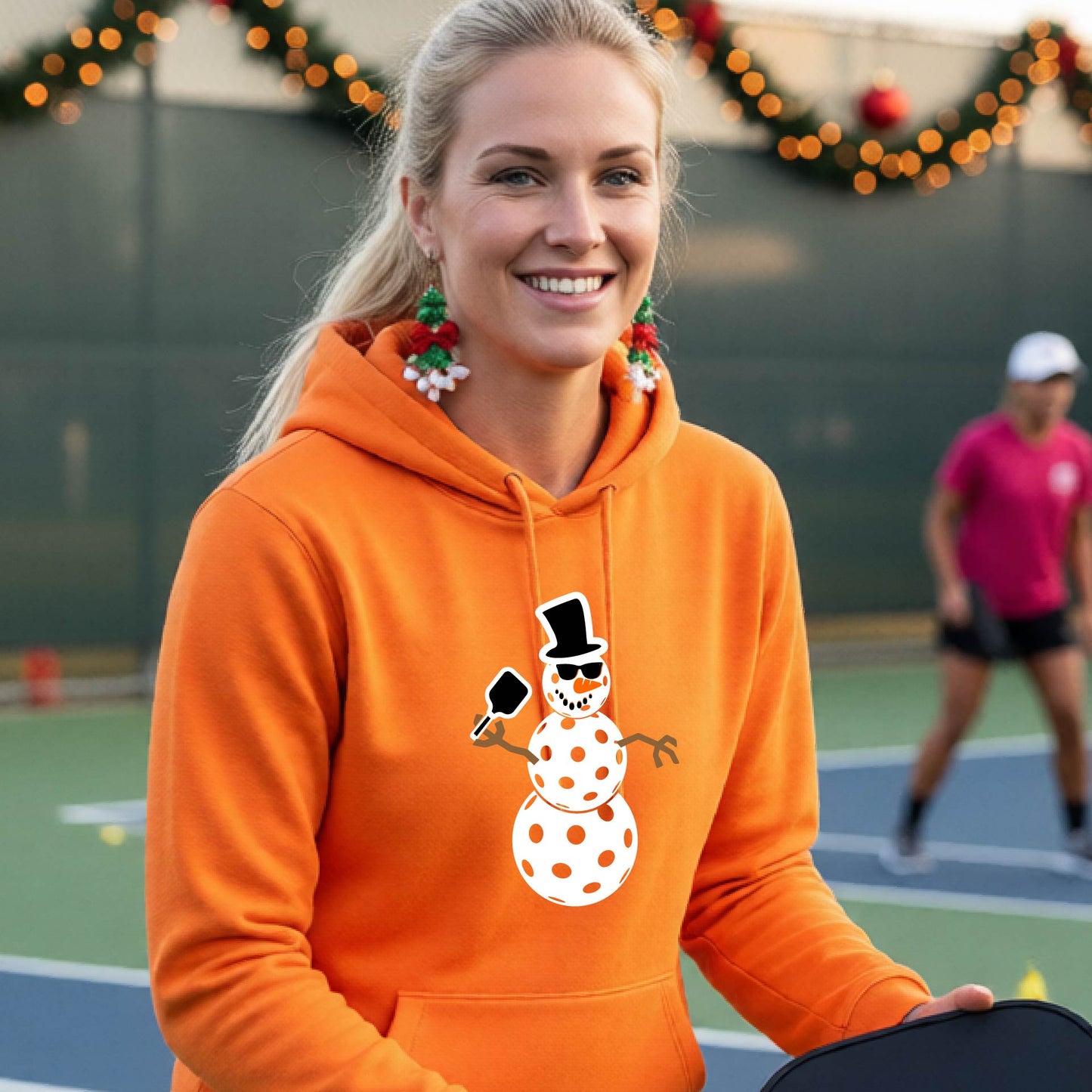 An man smiling on a picleball court wearing a bright orange hoodie with a white and blue pickleball snowman graphic on the front.
