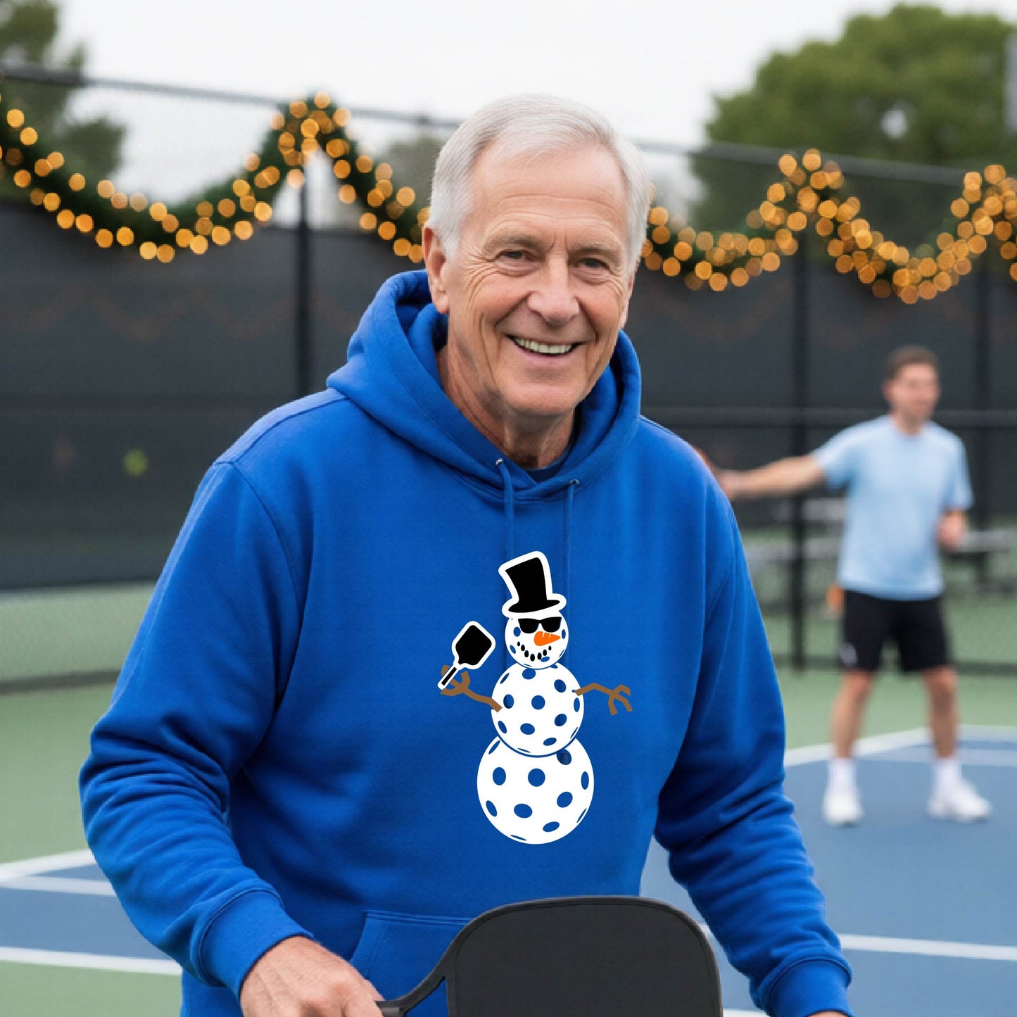 An man smiling on a pickleball court wearing a bright blue hoodie with a white and blue pickleball snowman graphic on the front.