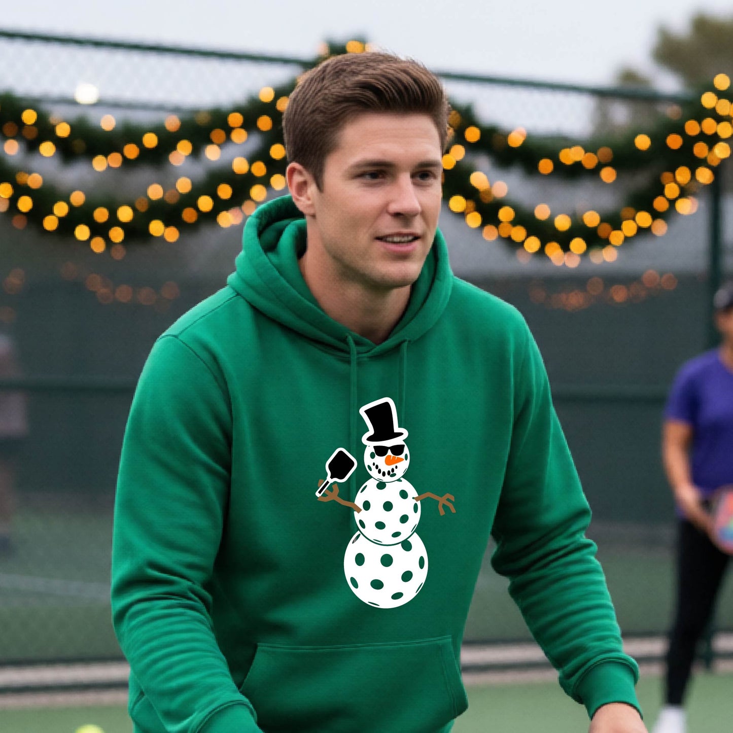 An man smiling on a picleball court wearing a bright green hoodie with a white and blue pickleball snowman graphic on the front.