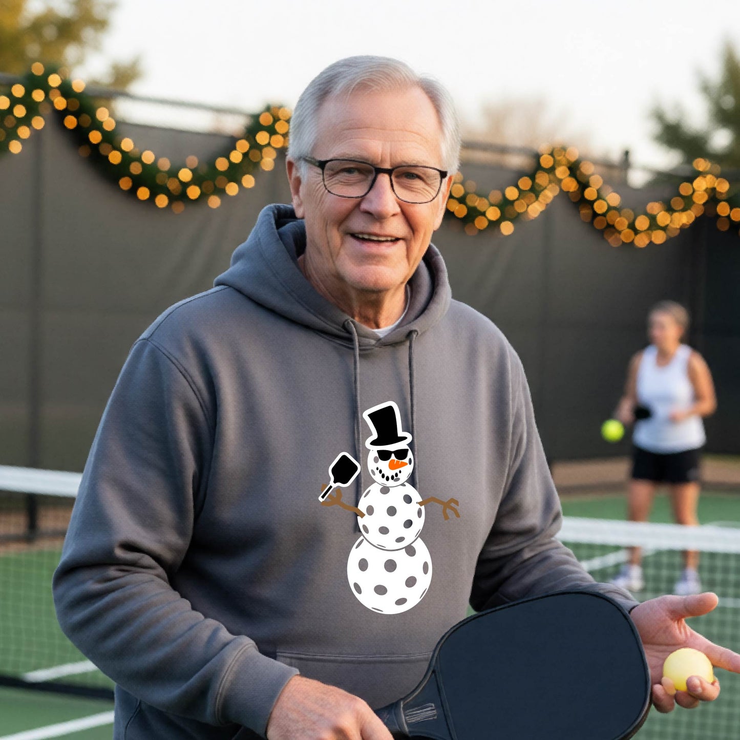 An man smiling on a picleball court wearing a gray hoodie with a white and blue pickleball snowman graphic on the front.
