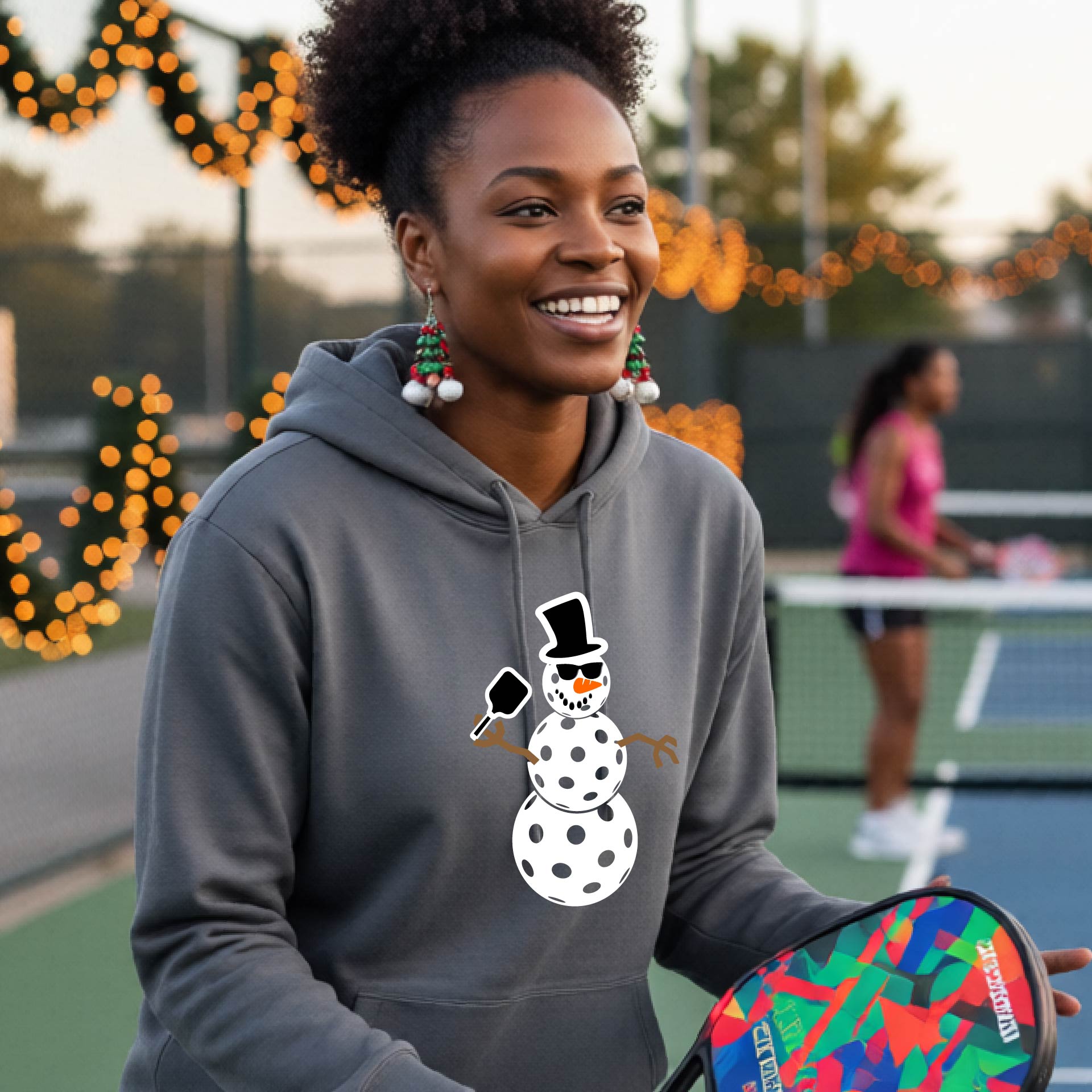 An woman smiling on a picleball court wearing a gray hoodie with a white and blue pickleball snowman graphic on the front.