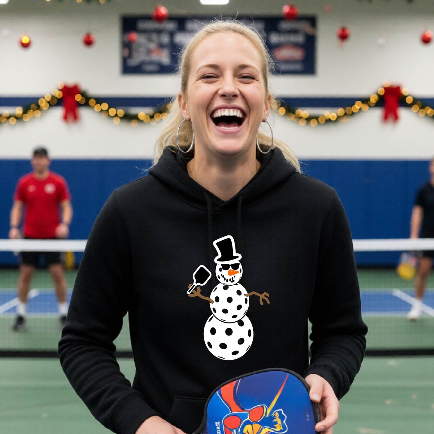 A black hooded sweatshirt featuring a pickleball graphic with a snowman made of pickleballs and a Christmas themed background. 