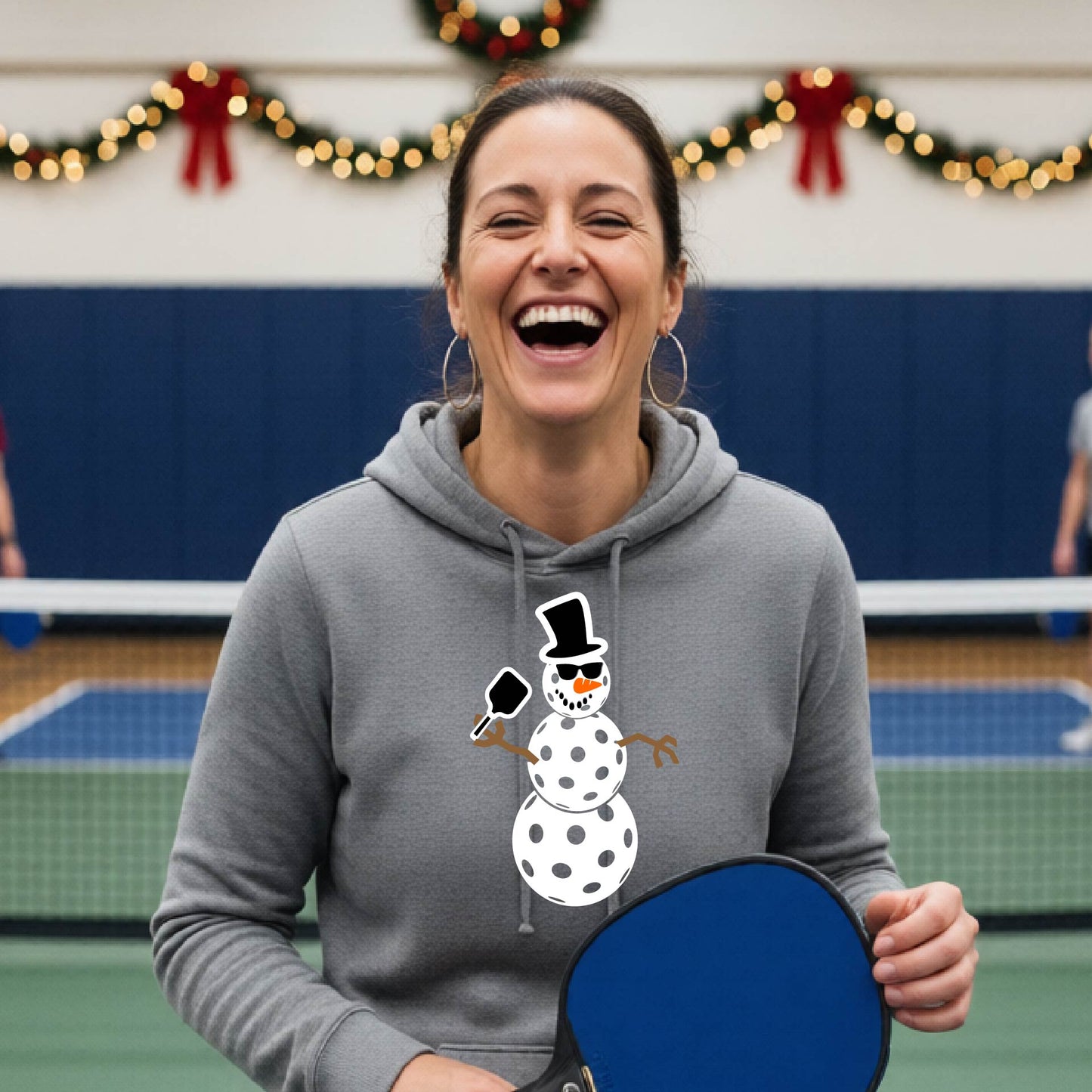 A grey hooded sweatshirt featuring a pickleball graphic with a snowman made of pickleballs and a Christmas themed background. 
