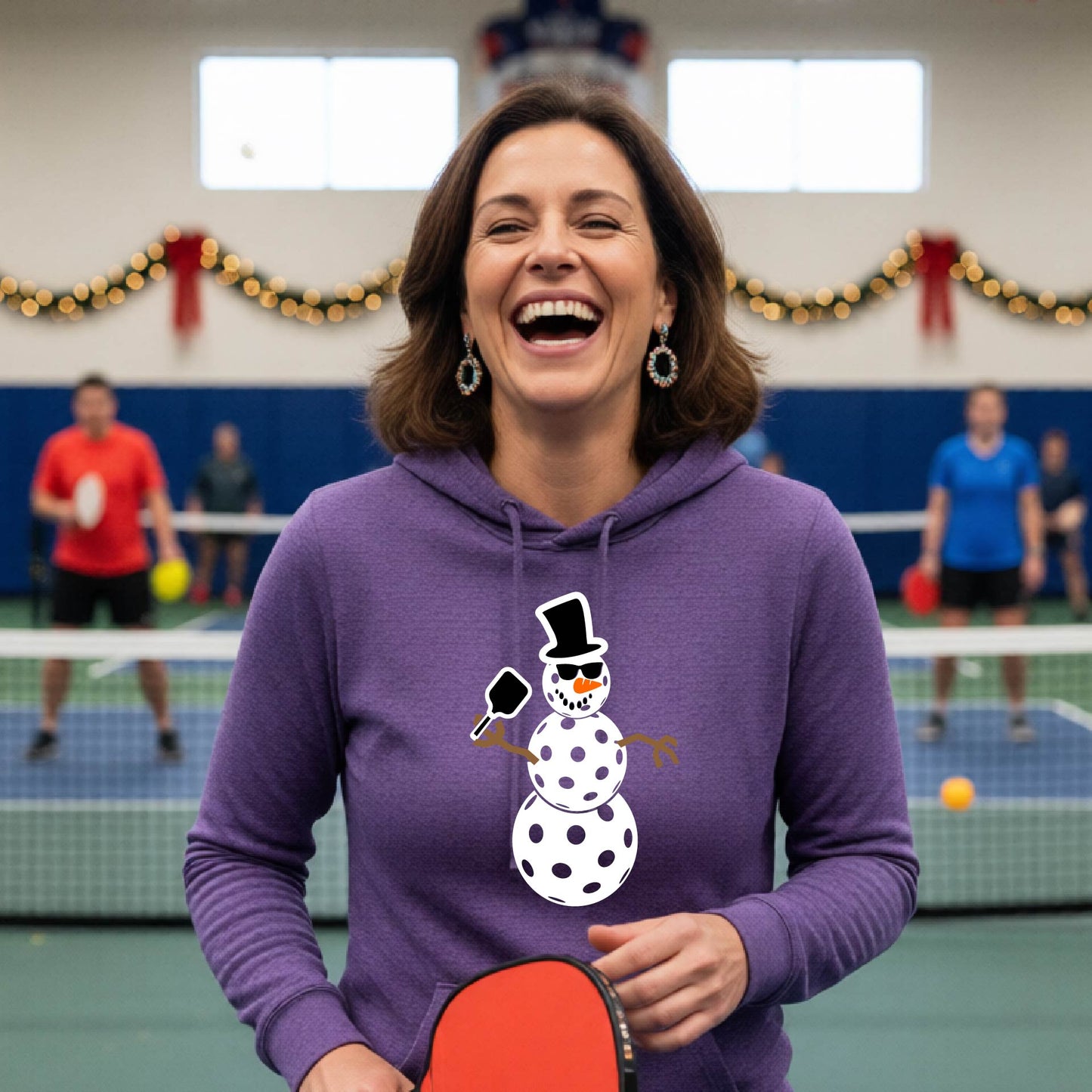A purple hooded sweatshirt featuring a pickleball graphic with a snowman made of pickleballs and a Christmas themed background. 