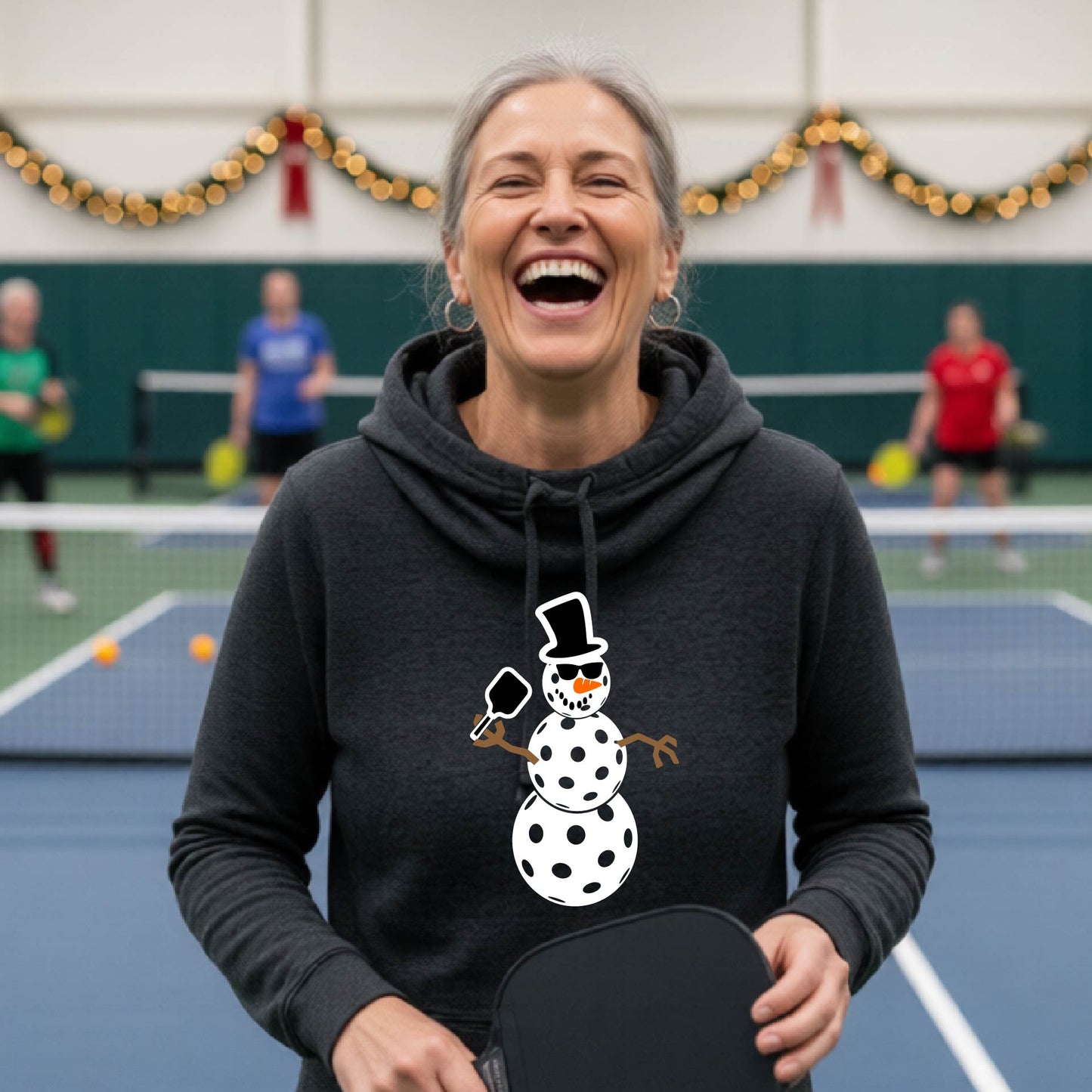 A heather black cowl-neck hooded sweatshirt featuring a pickleball graphic of a snowman made of white pickleballs and Christmas themed background. 