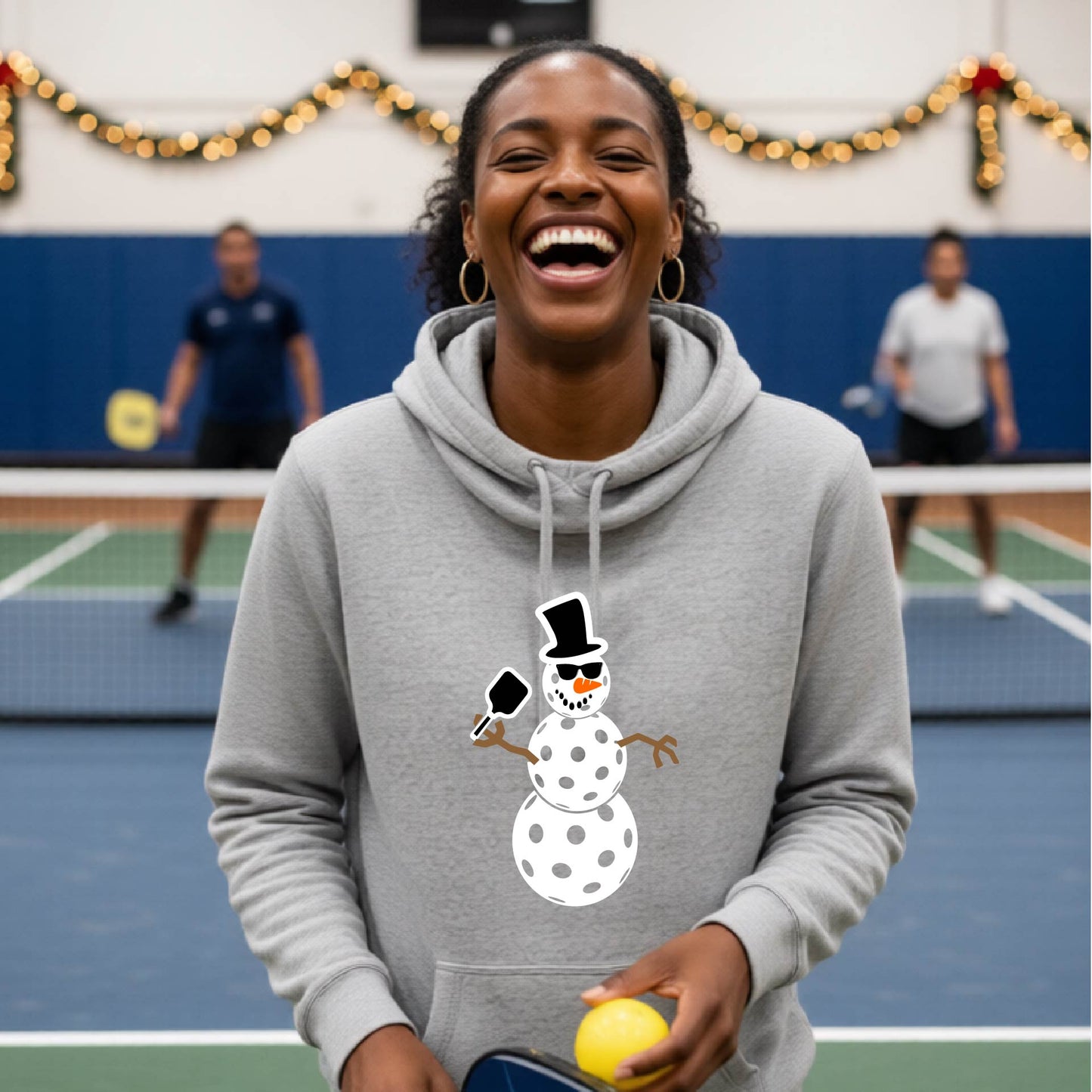 A gray cowl-neck hooded sweatshirt featuring a pickleball graphic of a snowman made of white pickleballs and Christmas themed background. 