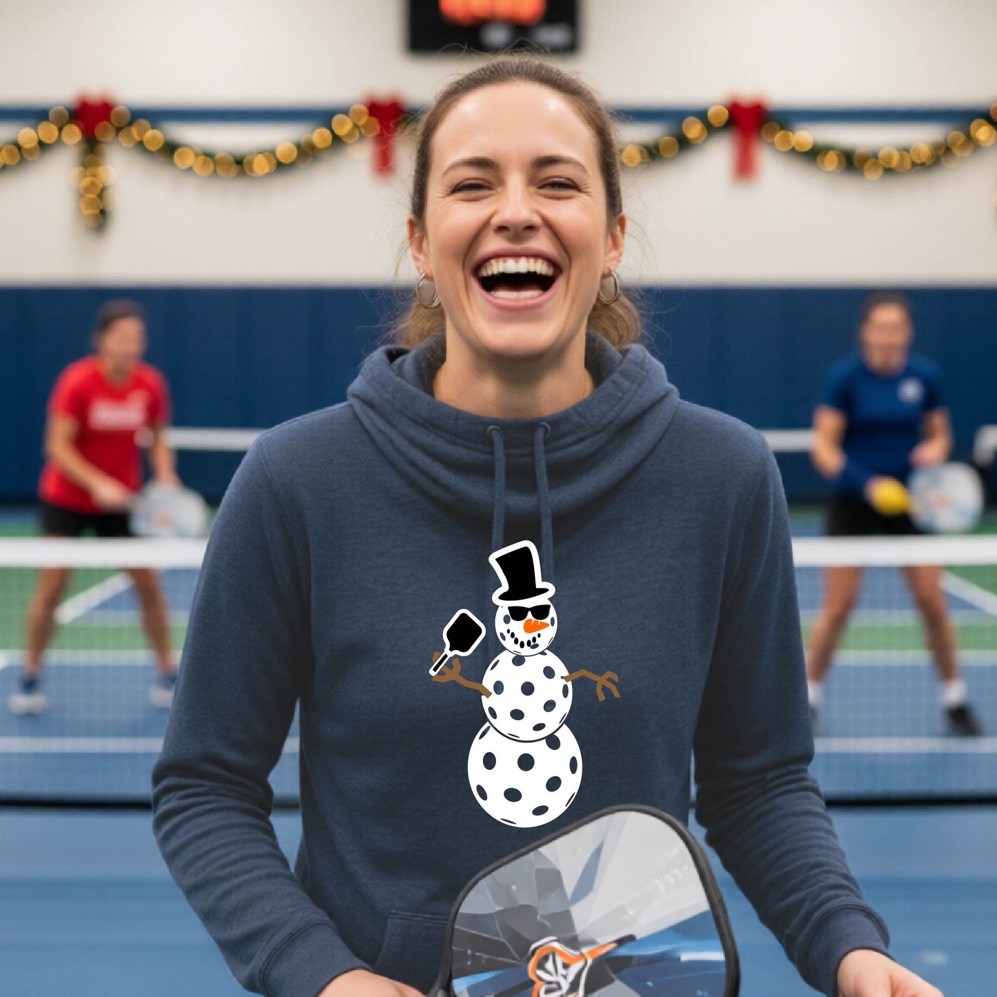 A heather navy cowl-neck hooded sweatshirt featuring a pickleball graphic of a snowman made of white pickleballs and Christmas themed background. 