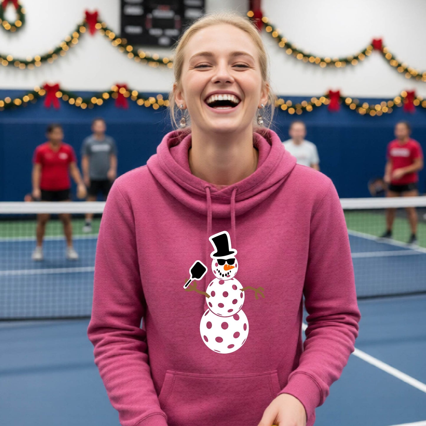 A pink cowl-neck hooded sweatshirt featuring a pickleball graphic of a snowman made of white pickleballs and Christmas themed background. 