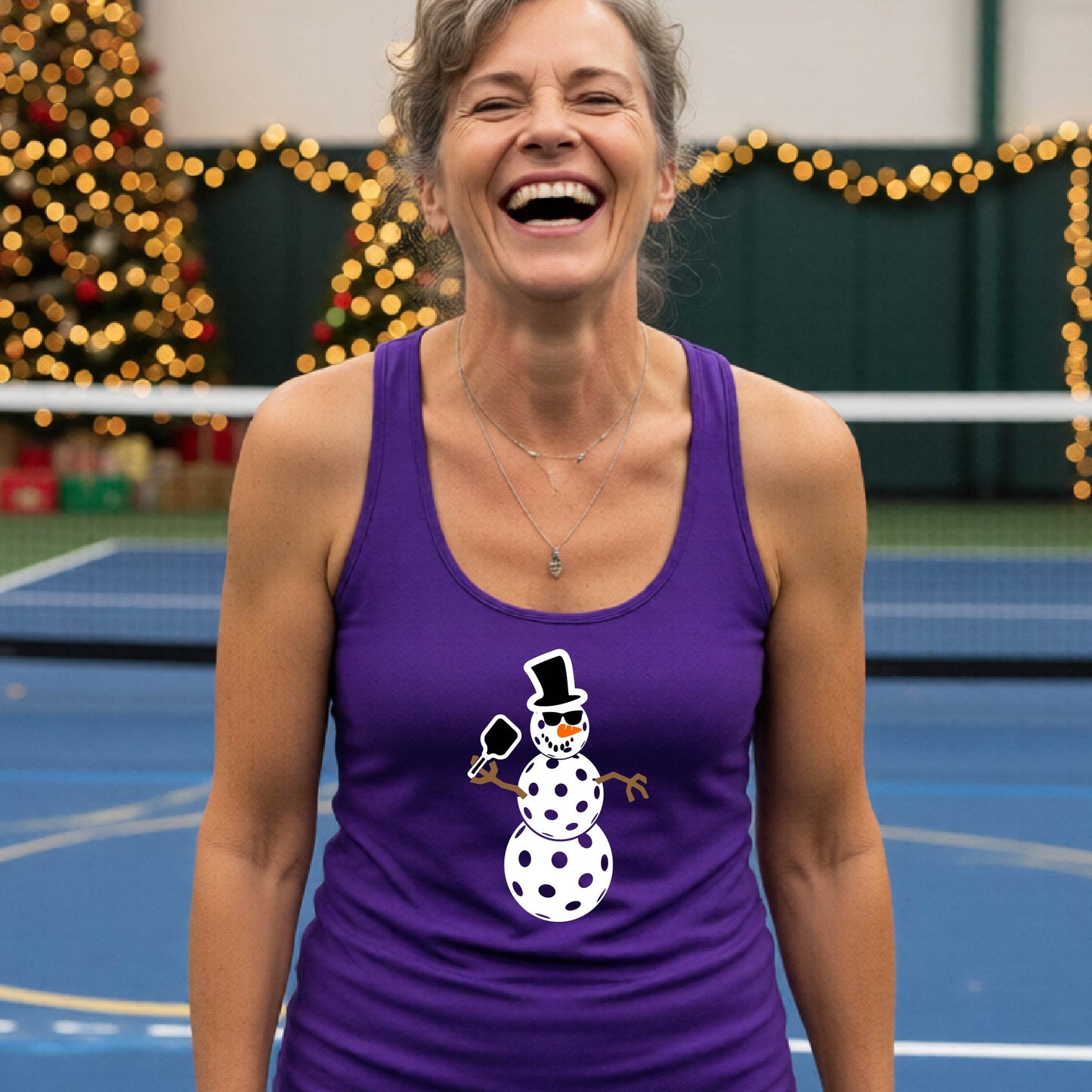 A woman wearing a purple racerback  tank top with a white and black pickleball snowman graphic, standing on a blue pickleball court with blurred Christmas decorations in the background.