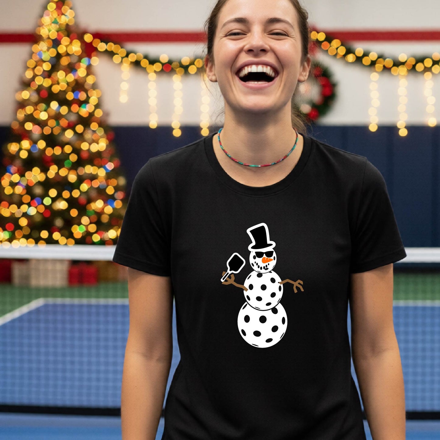 A woman wearing a black graphic t-shirt with a cartoon pickleball snowman wearing sunglasses and a top hat. The background shows a blurred indoor sports hall with holiday lights.