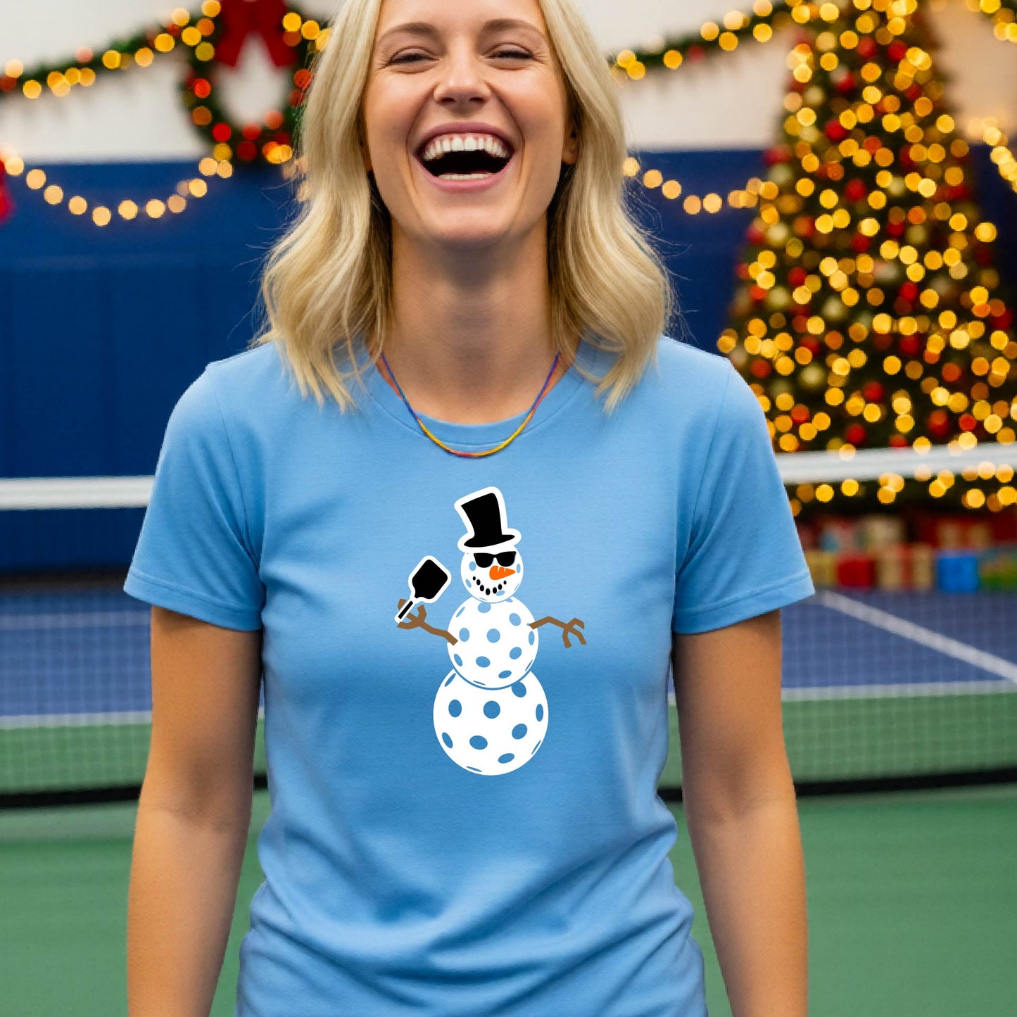 A smiling  woman wearing a light blue graphic t-shirt with a cartoon pickleball snowman wearing sunglasses and a top hat. The background shows a blurred indoor sports hall with holiday lights.