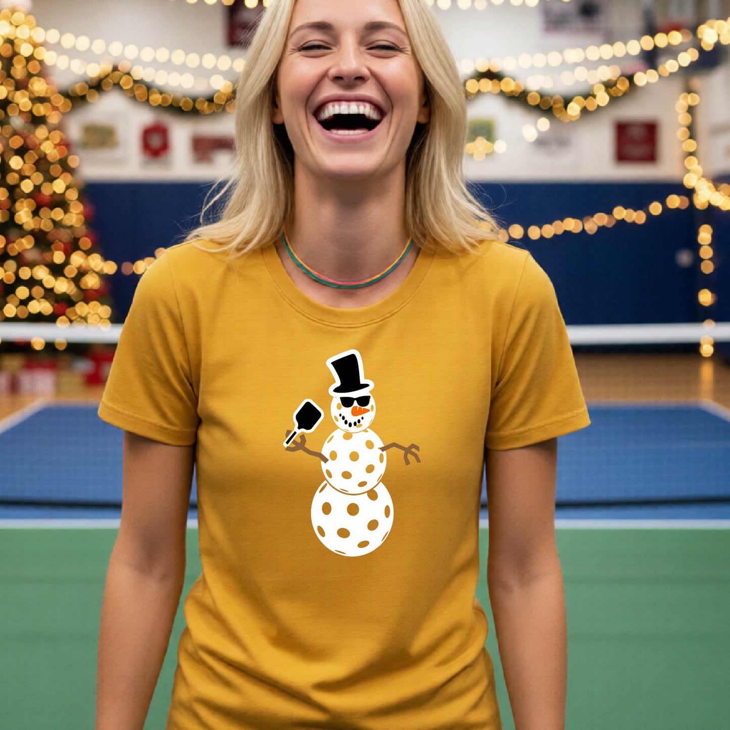 A smiling blonde woman wearing a mustard yellow gold graphic t-shirt with a cartoon pickleball snowman wearing sunglasses and a top hat. The background shows a blurred indoor sports hall with holiday lights.
