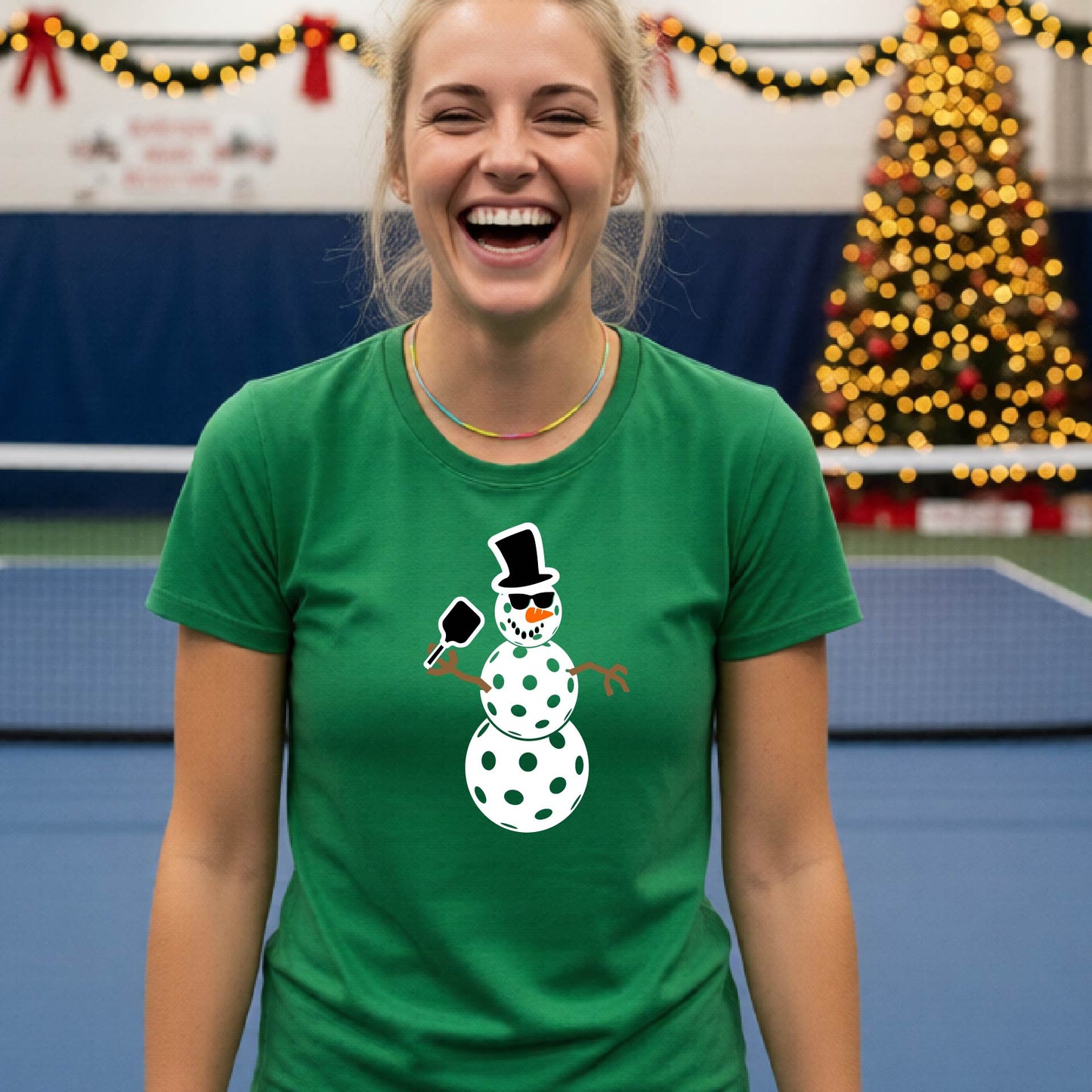 A smiling blonde woman wearing a green graphic t-shirt with a cartoon pickleball snowman wearing sunglasses and a top hat. The background shows a blurred indoor sports hall with holiday lights.
