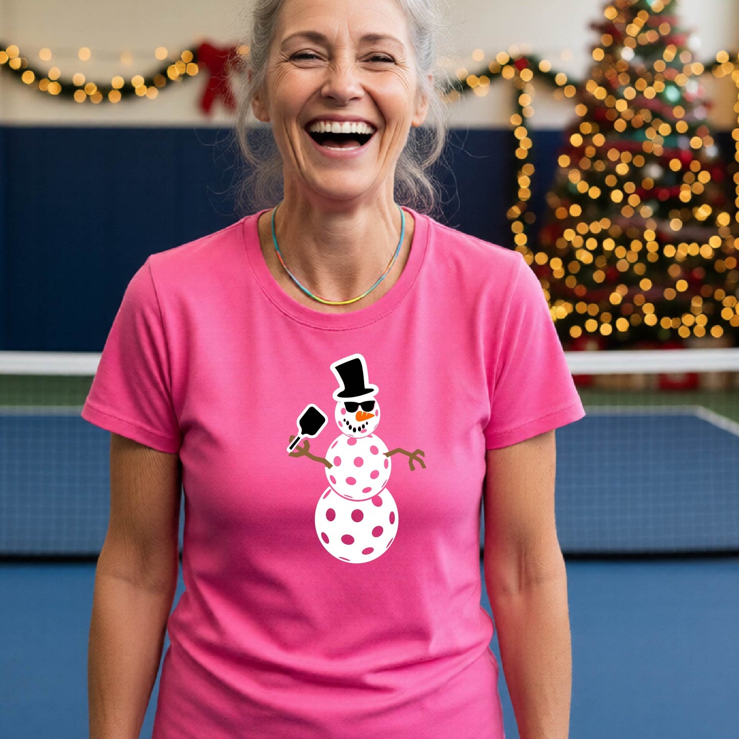 A smiling woman wearing a pink graphic t-shirt with a cartoon pickleball snowman wearing sunglasses and a top hat. The background shows a blurred indoor sports hall with holiday lights.