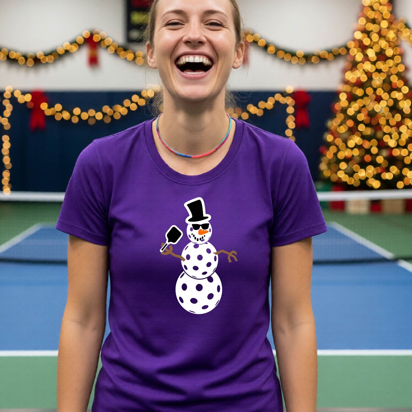 A smiling woman wearing a purple graphic t-shirt with a cartoon pickleball snowman wearing sunglasses and a top hat. The background shows a blurred indoor sports hall with holiday lights.