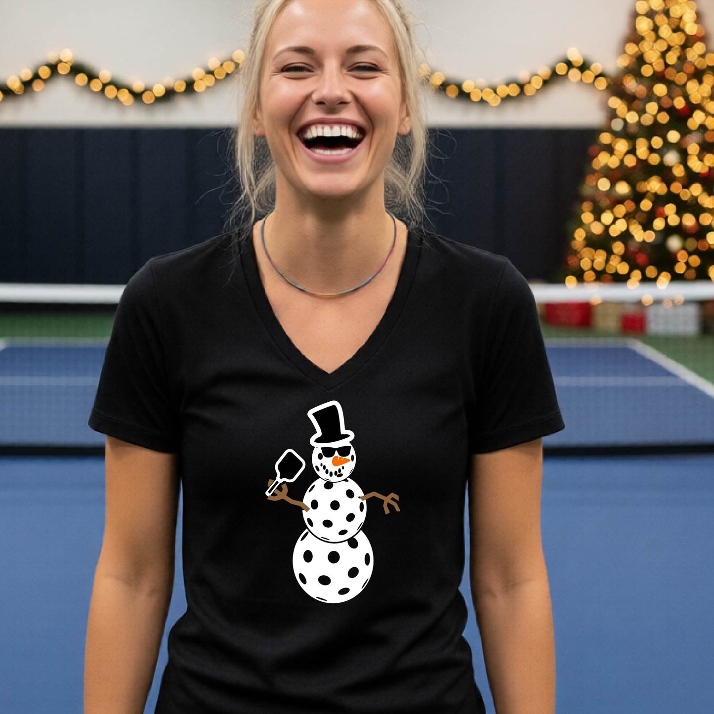 A woman wearing a black V-neck t-shirt with a cartoon snowman pickleball graphic, standing on a blue and green indoor court with a Christmas tree and holiday garlands in the background.