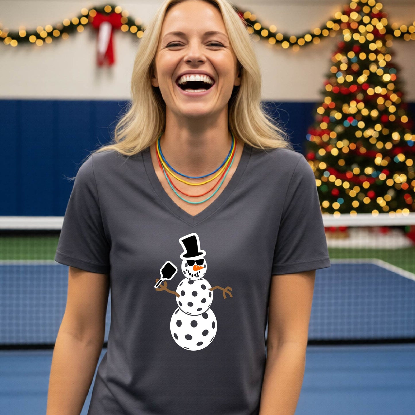 A woman wearing a dark gray V-neck t-shirt with a cartoon snowman pickleball graphic, standing on a blue and green indoor court with a Christmas tree and holiday garlands in the background.