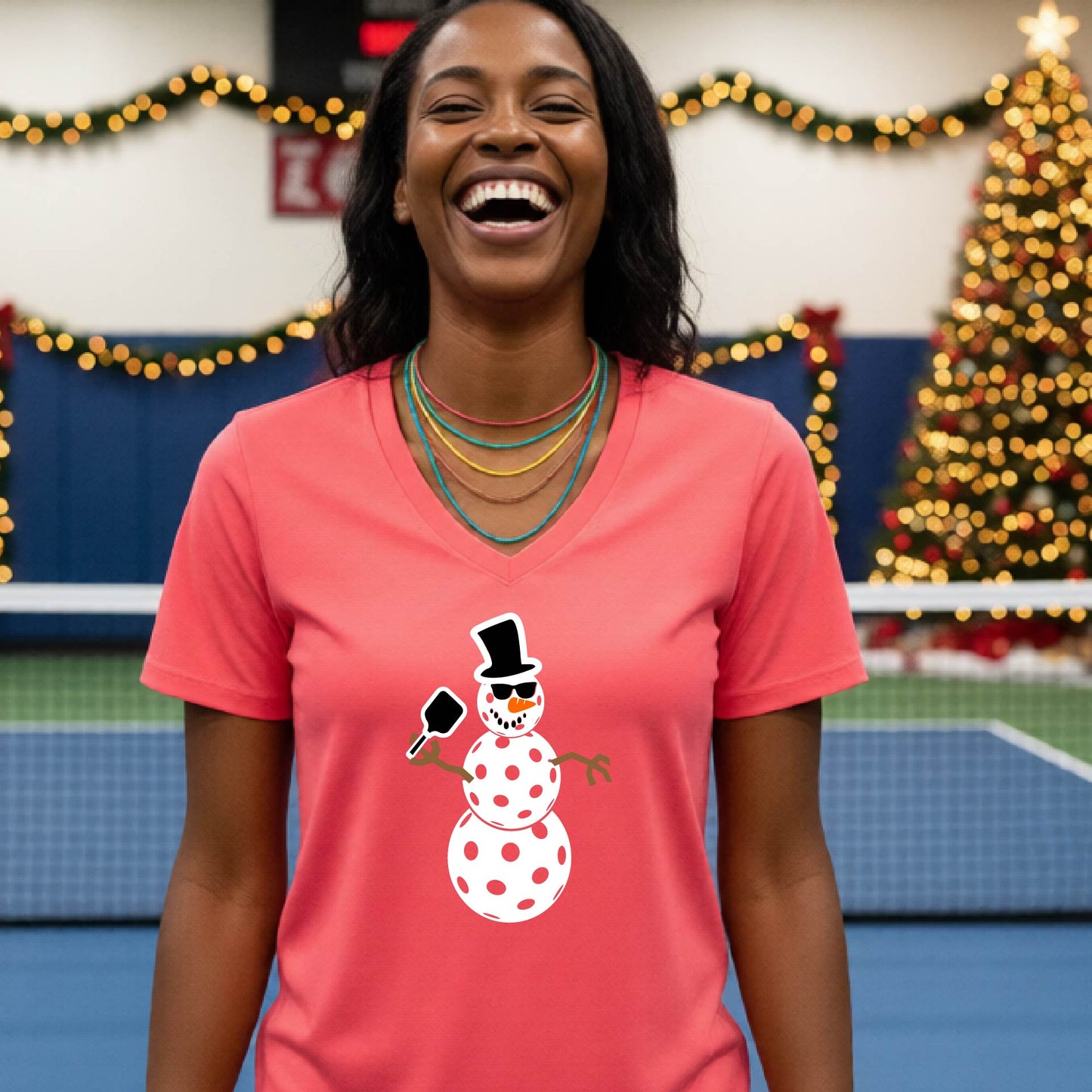 A woman wearing a bright coral-pink V-neck t-shirt with a cartoon snowman pickleball graphic, standing on a blue and green indoor court with a Christmas tree and holiday garlands in the background.