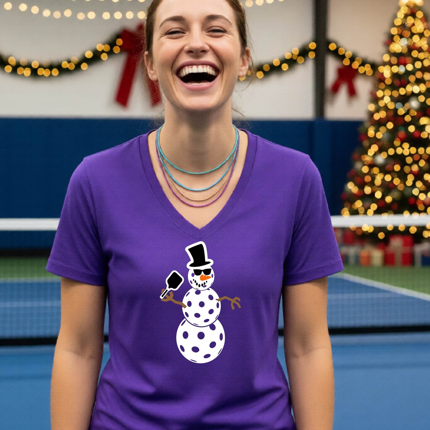 A woman wearing a bright purple V-neck t-shirt with a cartoon snowman pickleball graphic, standing on a blue and green indoor court with a Christmas tree and holiday garlands in the background.