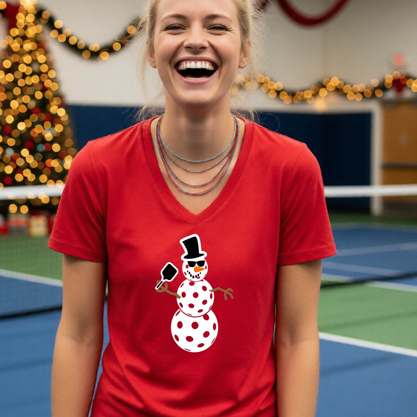 A woman wearing a bright red V-neck t-shirt with a cartoon snowman pickleball graphic, standing on a blue and green indoor court with a Christmas tree and holiday garlands in the background.