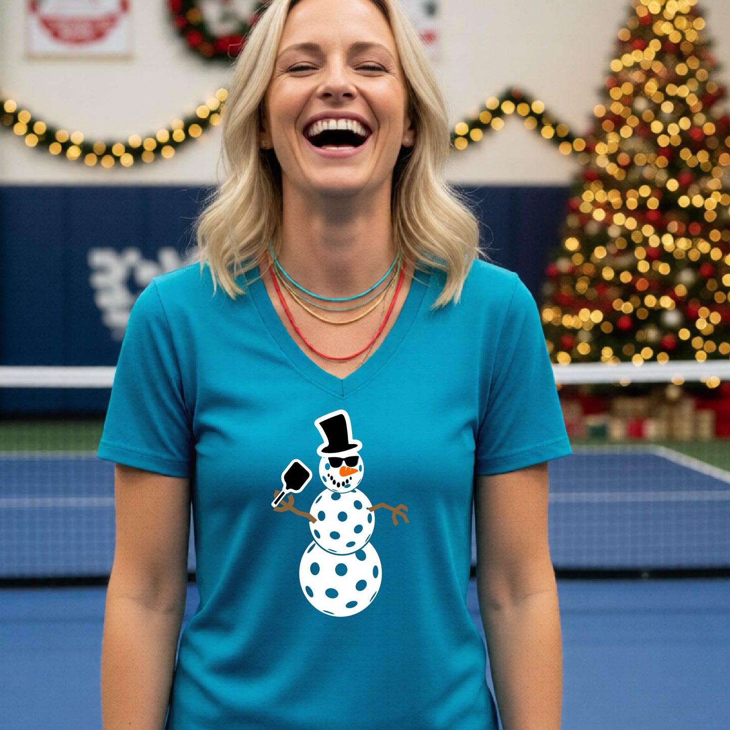 A woman wearing a bright teal V-neck t-shirt with a cartoon snowman pickleball graphic, standing on a blue and green indoor court with a Christmas tree and holiday garlands in the background.