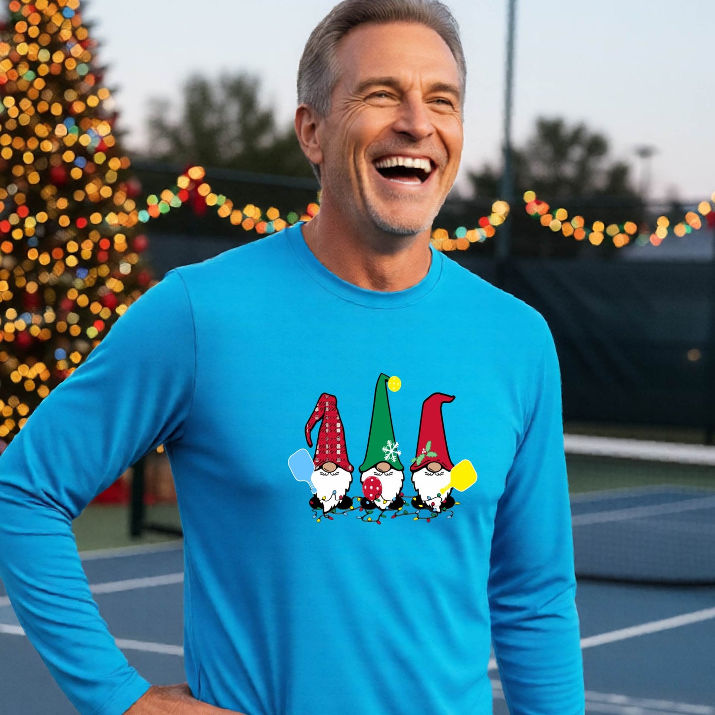 A man laughing on an outdoor pickleball court at dusk, wearing a bright blue long-sleeve shirt with three colorful gnome holding a pickleball paddles graphic on the chest. The background features warm string lights and a decorated Christmas tree.