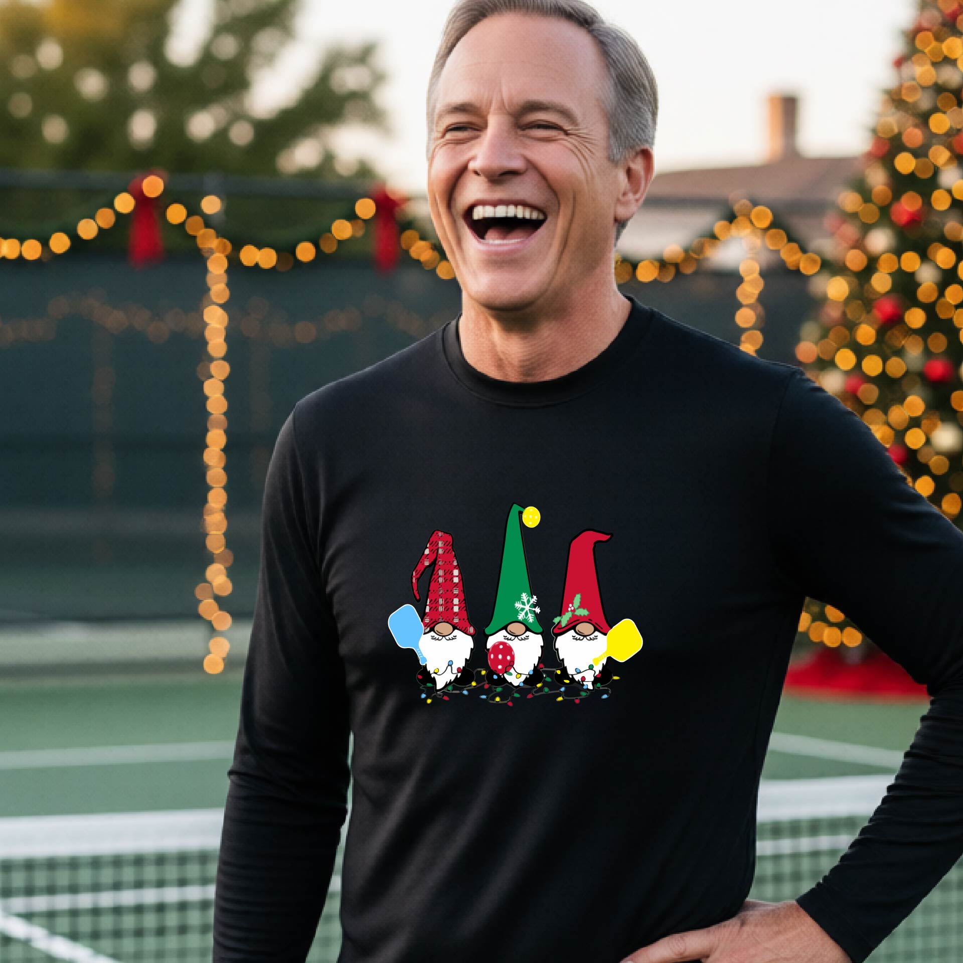 A man laughing on an outdoor pickleball court at dusk, wearing a black long-sleeve shirt with three colorful gnome holding a pickleball paddles graphic on the chest. The background features warm string lights and a decorated Christmas tree.