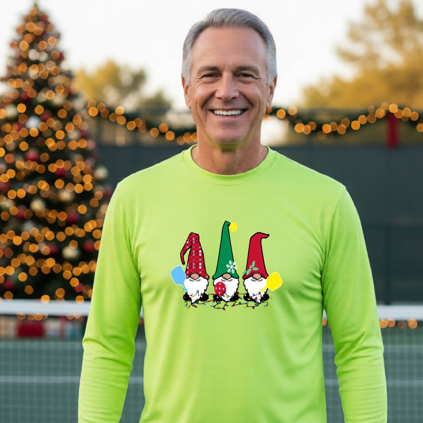 A man laughing on an outdoor pickleball court at dusk, wearing a lime green long-sleeve shirt with three colorful gnome holding a pickleball paddles graphic on the chest. The background features warm string lights and a decorated Christmas tree.