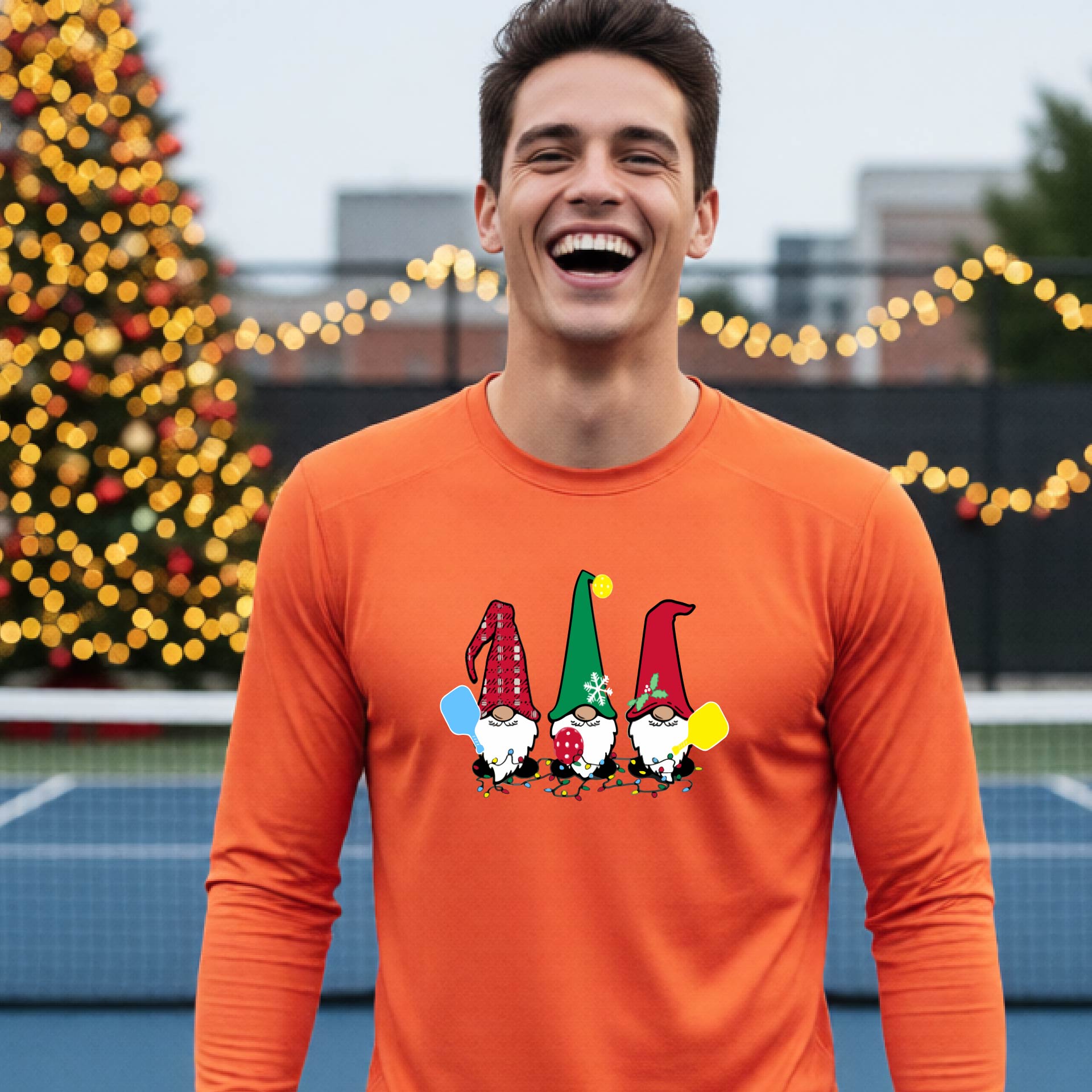 A man laughing on an outdoor pickleball court at dusk, wearing a bright orange long-sleeve shirt with three colorful gnome holding a pickleball paddles graphic on the chest. The background features warm string lights and a decorated Christmas tree.