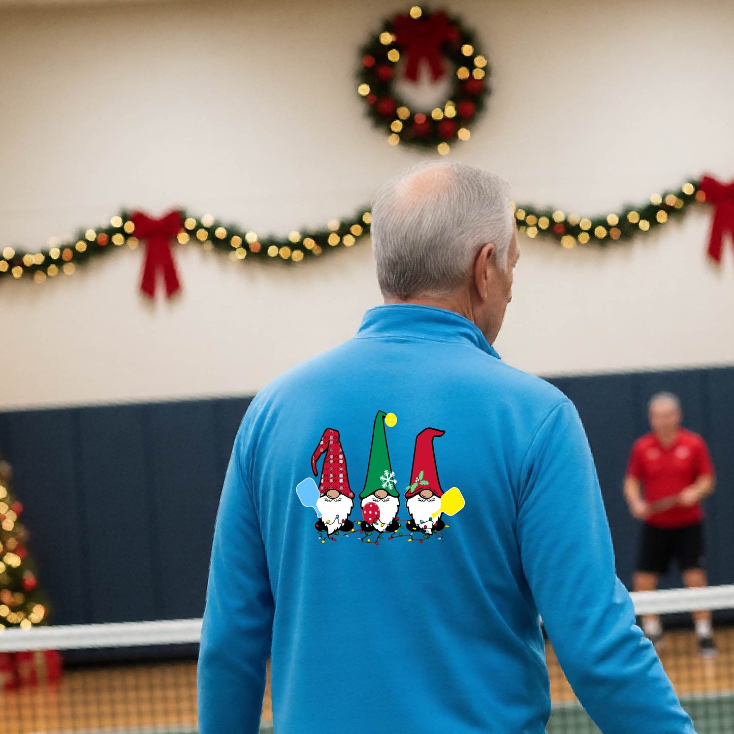 A man viewed from behind wearing a light blue long-sleeve quarter-zip pullover with a graphic of three Christmas gnomes on the back holding pickleball paddles, standing on pickleball court with holiday garlands in the background.