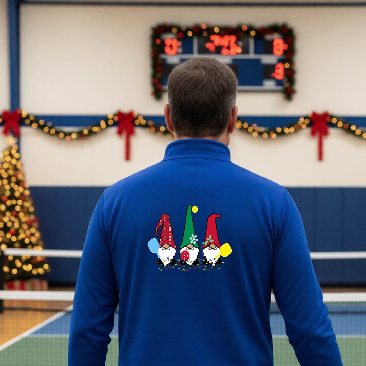 A man viewed from behind wearing a royal blue long-sleeve quarter-zip pullover with a graphic of three Christmas gnomes on the back holding pickleball paddles, standing on pickleball court with holiday garlands in the background.