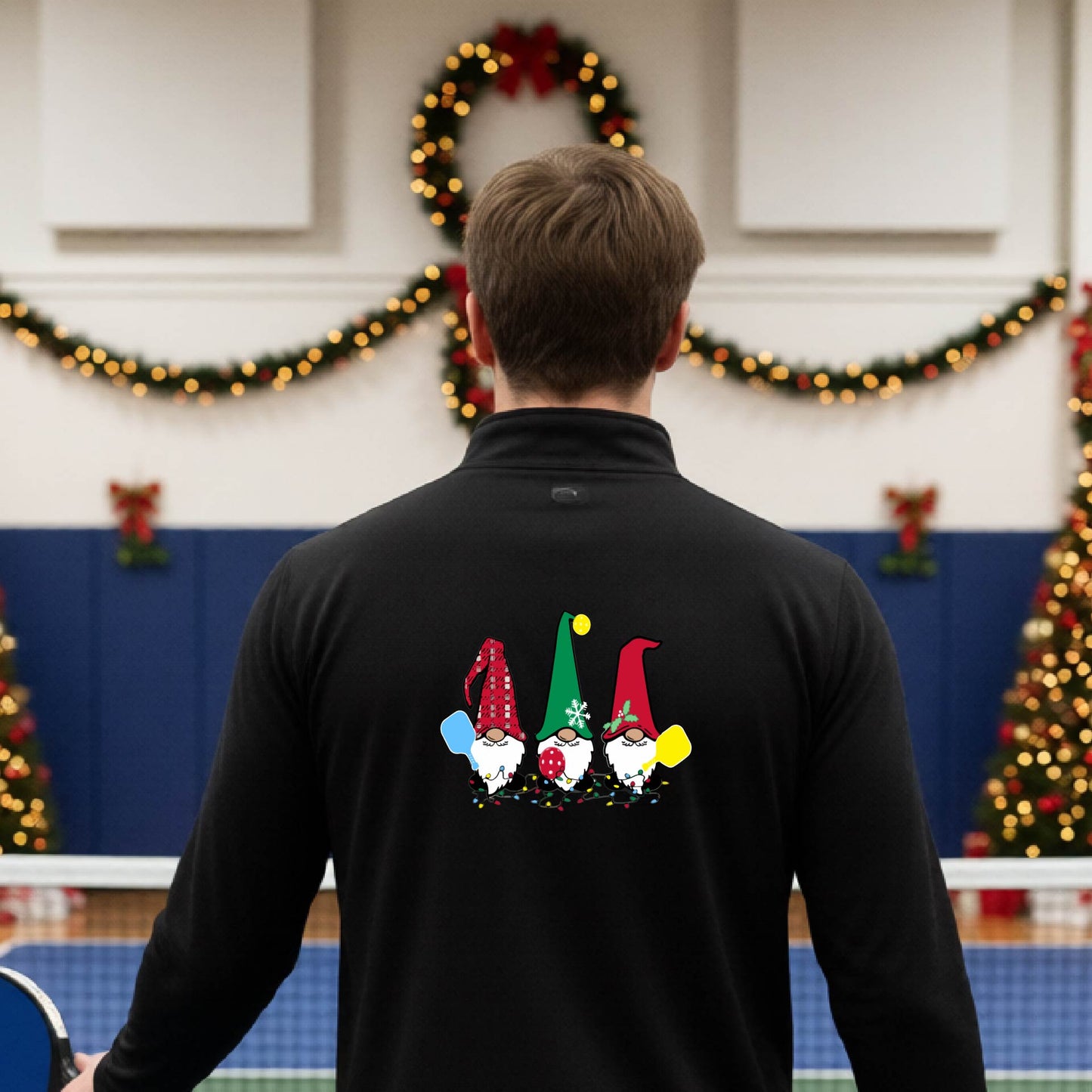 A man viewed from behind wearing a black long-sleeve quarter-zip pullover with a graphic of three Christmas gnomes on the back holding pickleball paddles, standing on pickleball court with holiday garlands in the background.