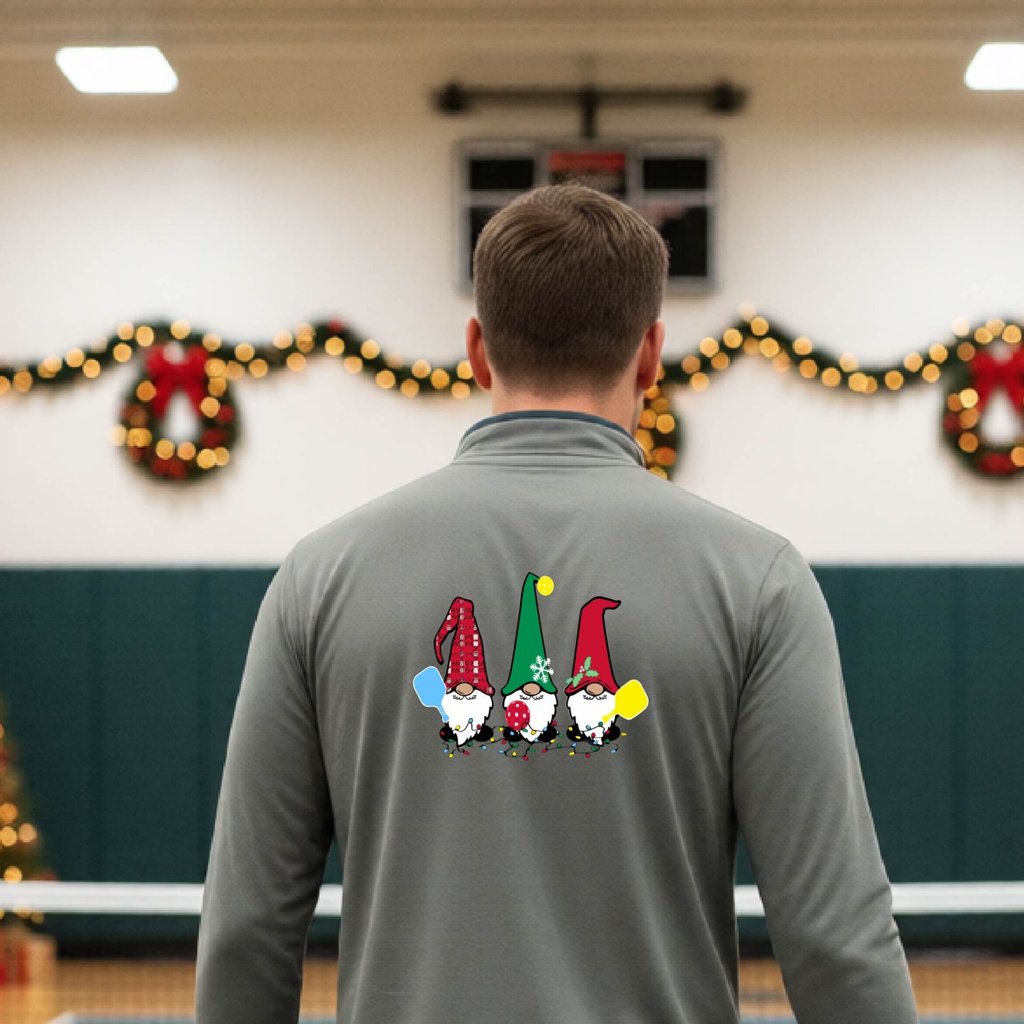 A man viewed from behind wearing a gray long-sleeve quarter-zip pullover with a graphic of three Christmas gnomes on the back holding pickleball paddles, standing on pickleball court with holiday garlands in the background.